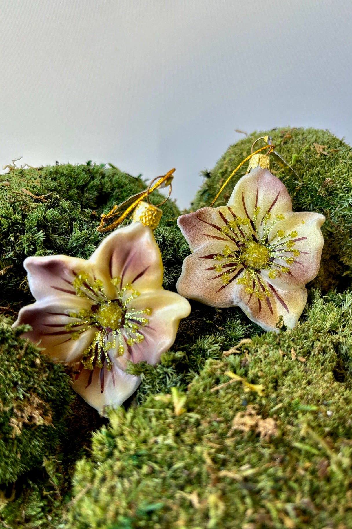 Two white glass ornaments with splashes of purple on the petals with pale green glitter displayed on a moss mound against a white wall. ©Sprout Home