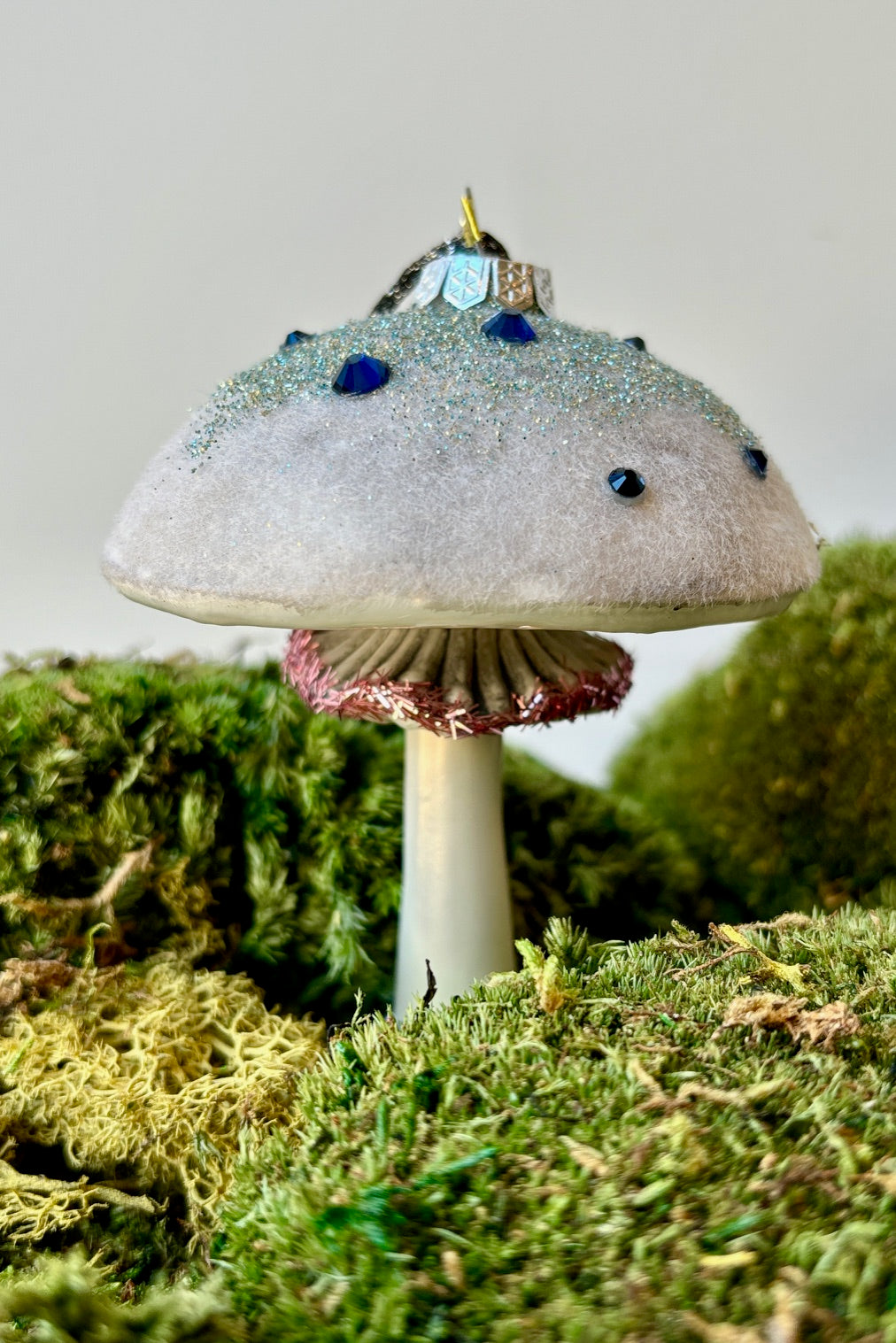 Frostfield mushroom in white glass with an opalescent glitter frosted cap accented with blue gemstones and a small ruffled underneath outlined in pink shredded glitter on a mound of moss. ©Sprout Home