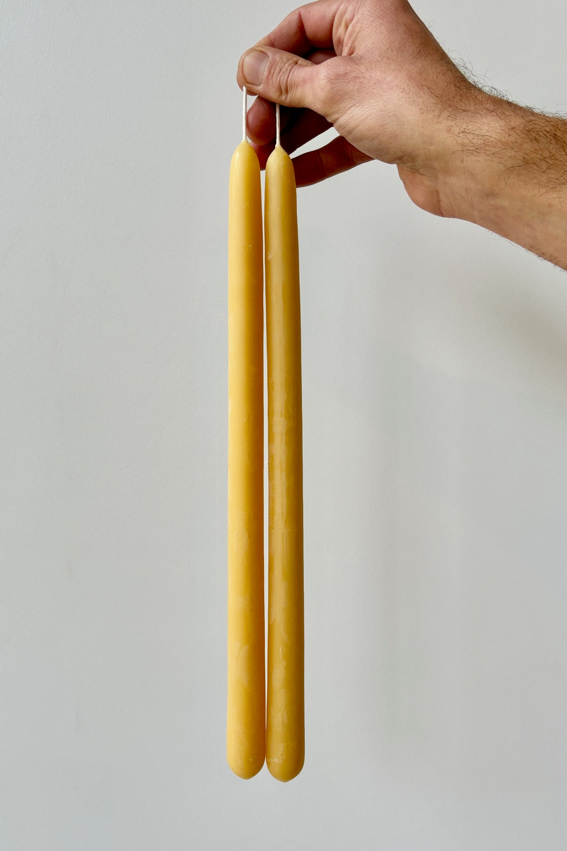 Hand holding a pair of natural beeswax taper candles against a white background.