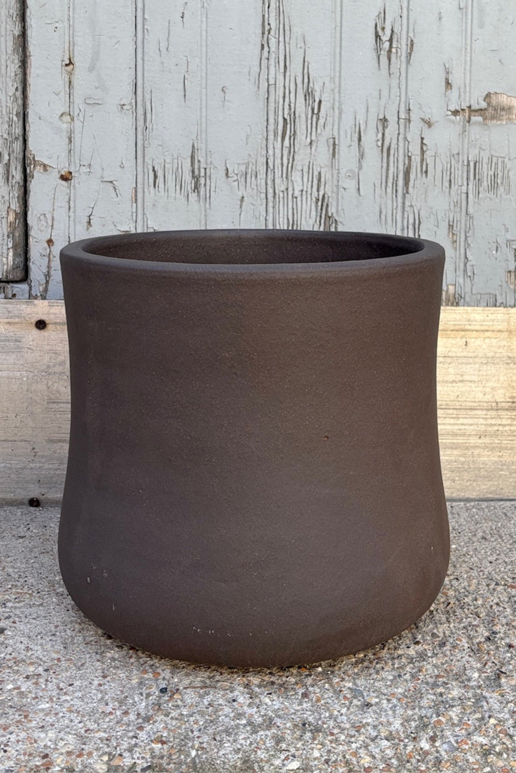 Medium Brown Terra cotta pot with an hourglass shape against a grey wall on a cement surface. Planter contains a drainage hole.