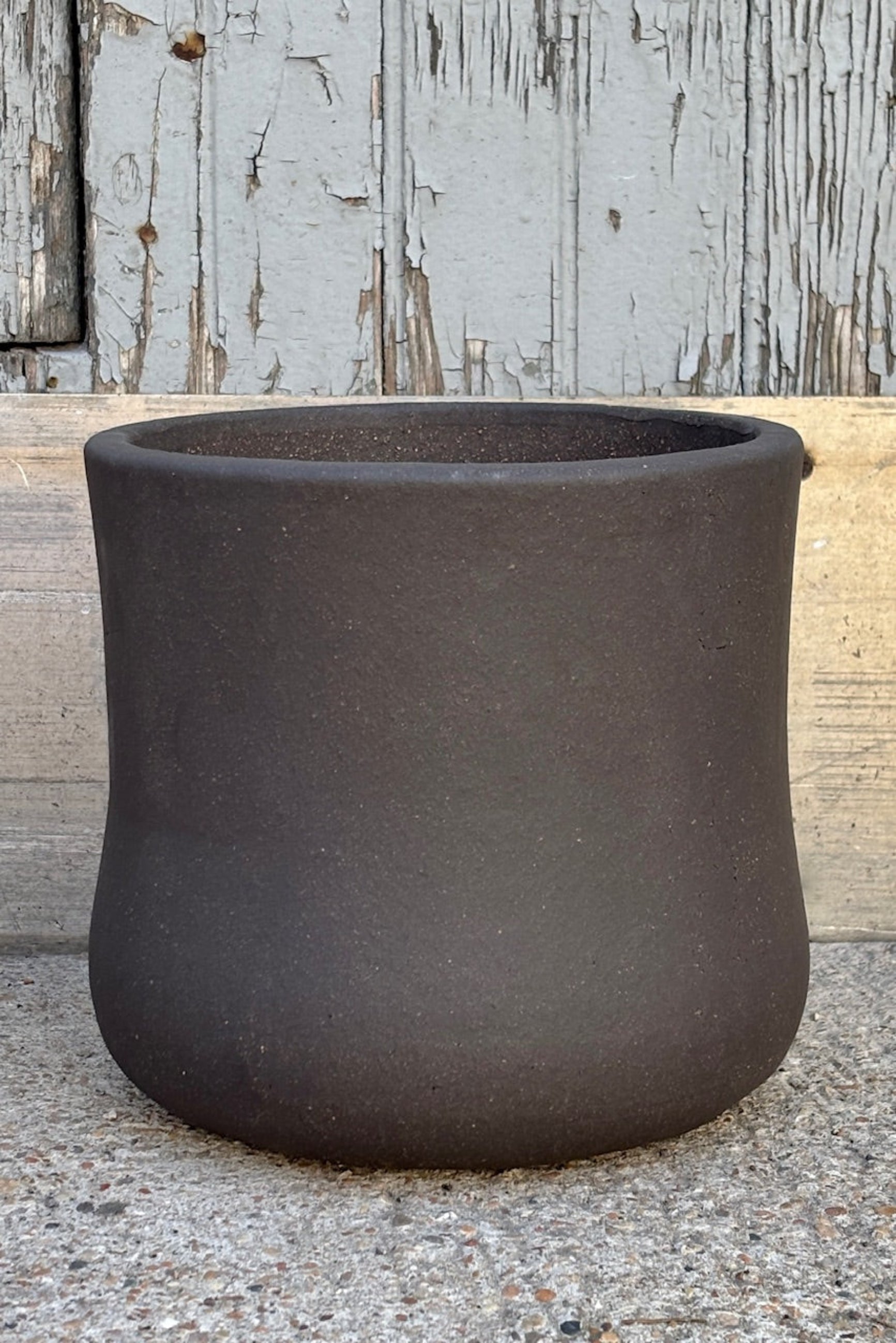 Small Brown Terra cotta pot with an hourglass shape against a grey wall on a cement surface. Planter contains a drainage hole.