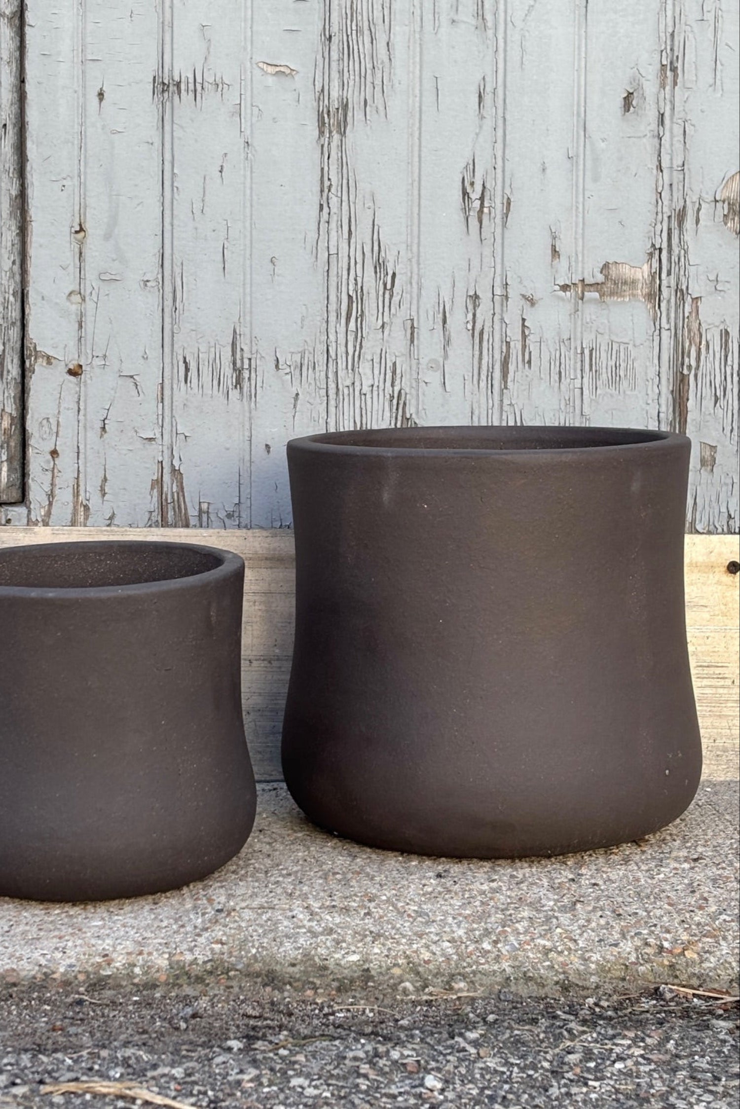 Medium  and small Brown Terra cotta pots with hourglass shape against a grey wall on a cement surface. Planters contain a drainage hole.  ©Sprout Home