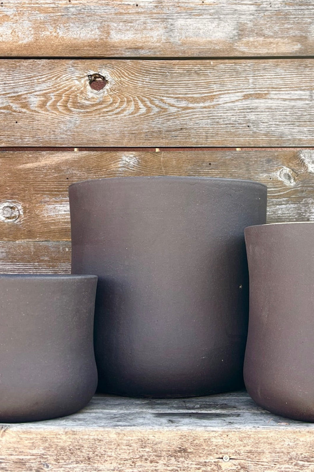 Trio of Brown Terra cotta pots with an hourglass shape against a wood wall. Planters contain a drainage hole.  ©Sprout Home