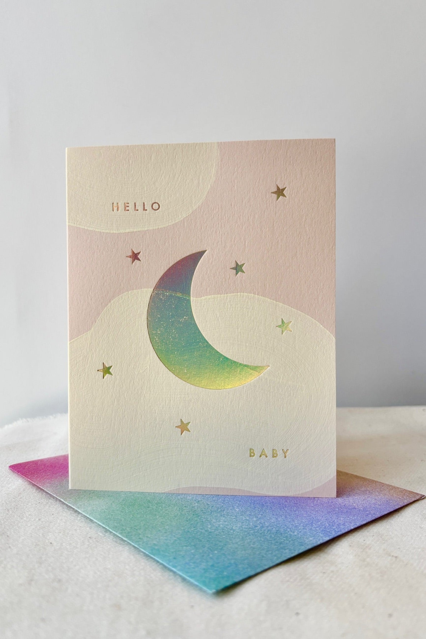 The Moon Card by Moglea showing the front of the card with the text Hello Baby in gold lettering and an envelope with airbrushed pastel colors against a white wall