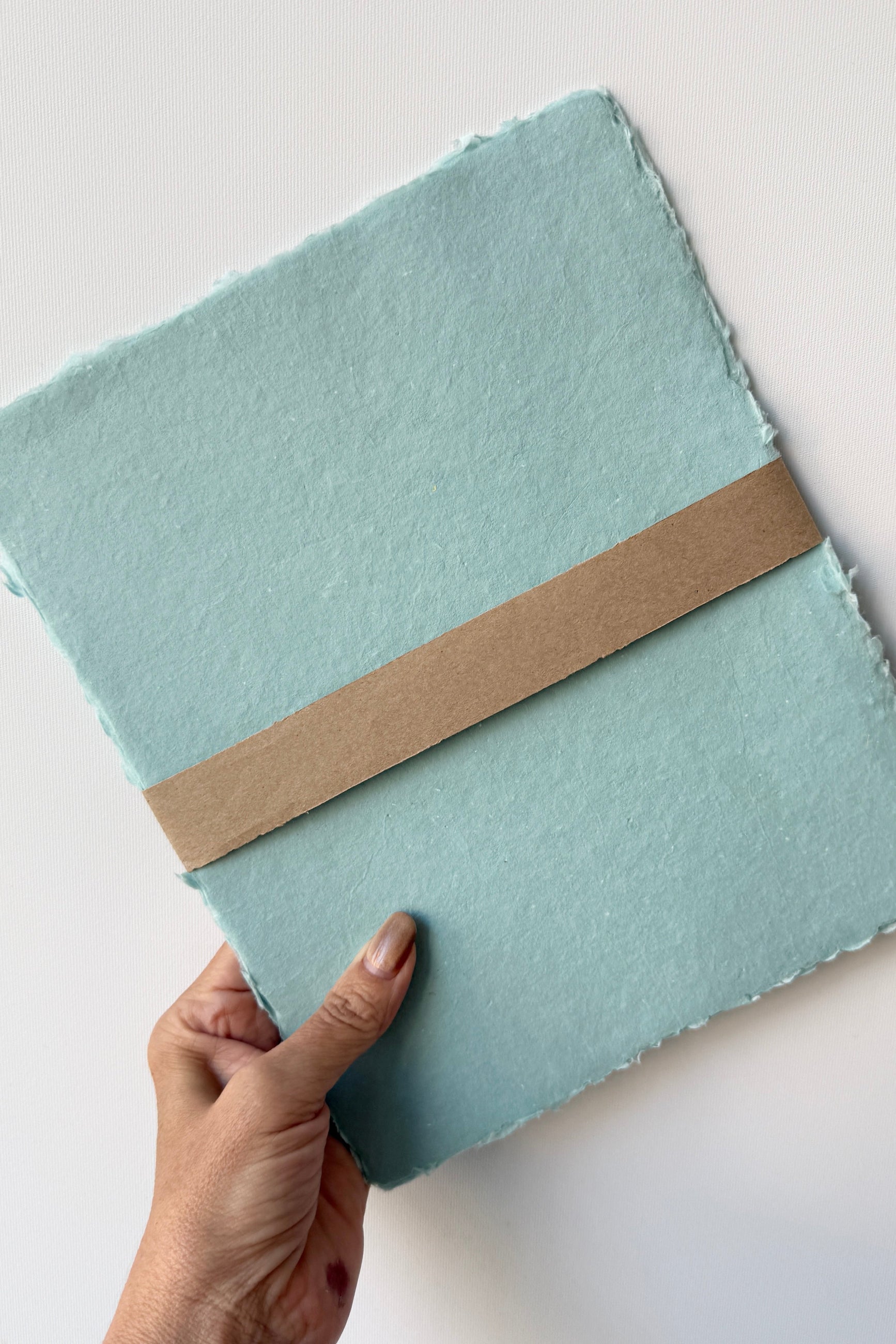 Light blue Handmade Celadon Paper Sheet Set in 8x10 by Sustainable Paper +Craft held by a hand on a light gray background ©Sprout Hme