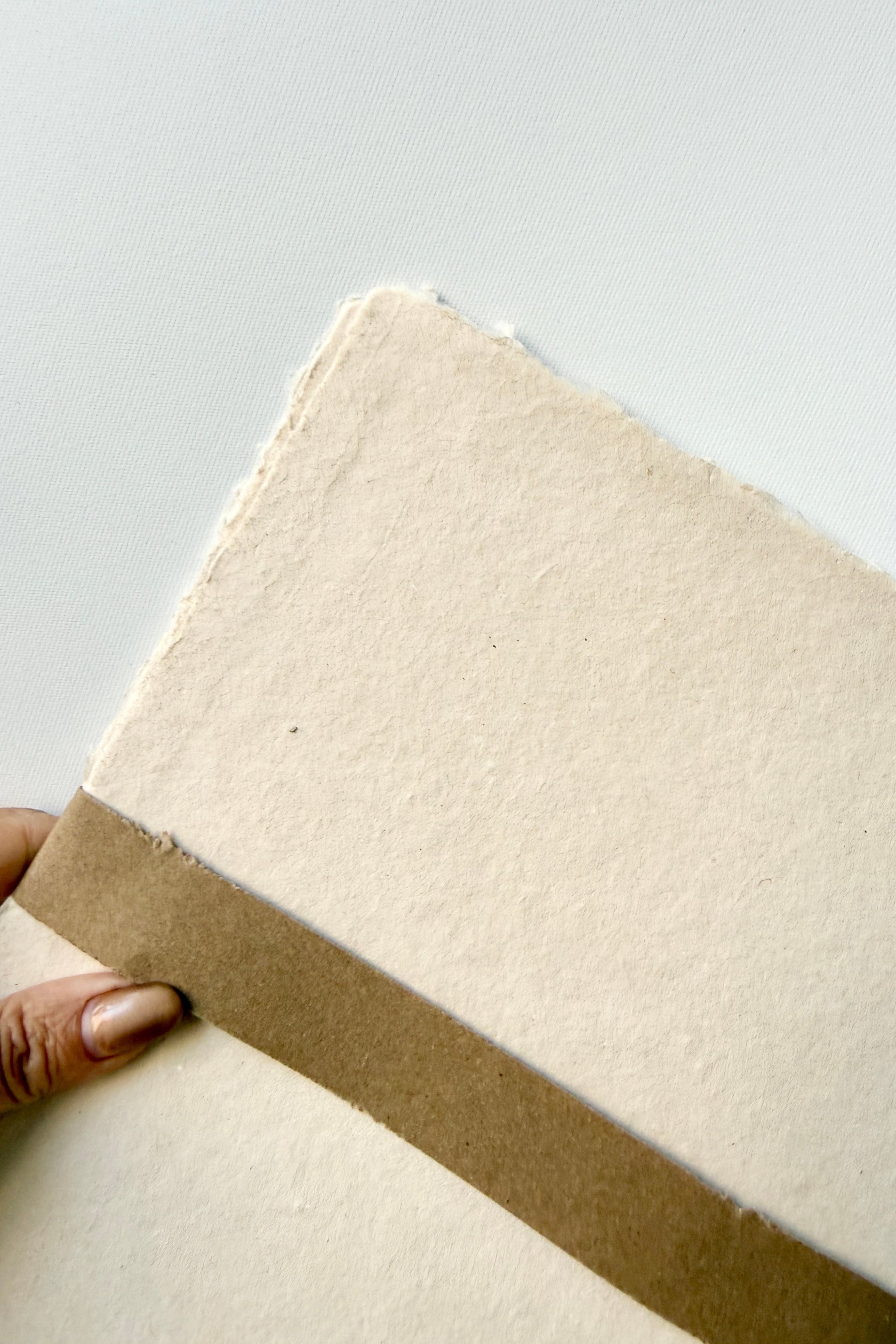 Hand holding a Handmade Buff Paper Sheet Set in 8x10 with a brown band by Sustainable Paper & Craft against a light white background ©Sprout Home