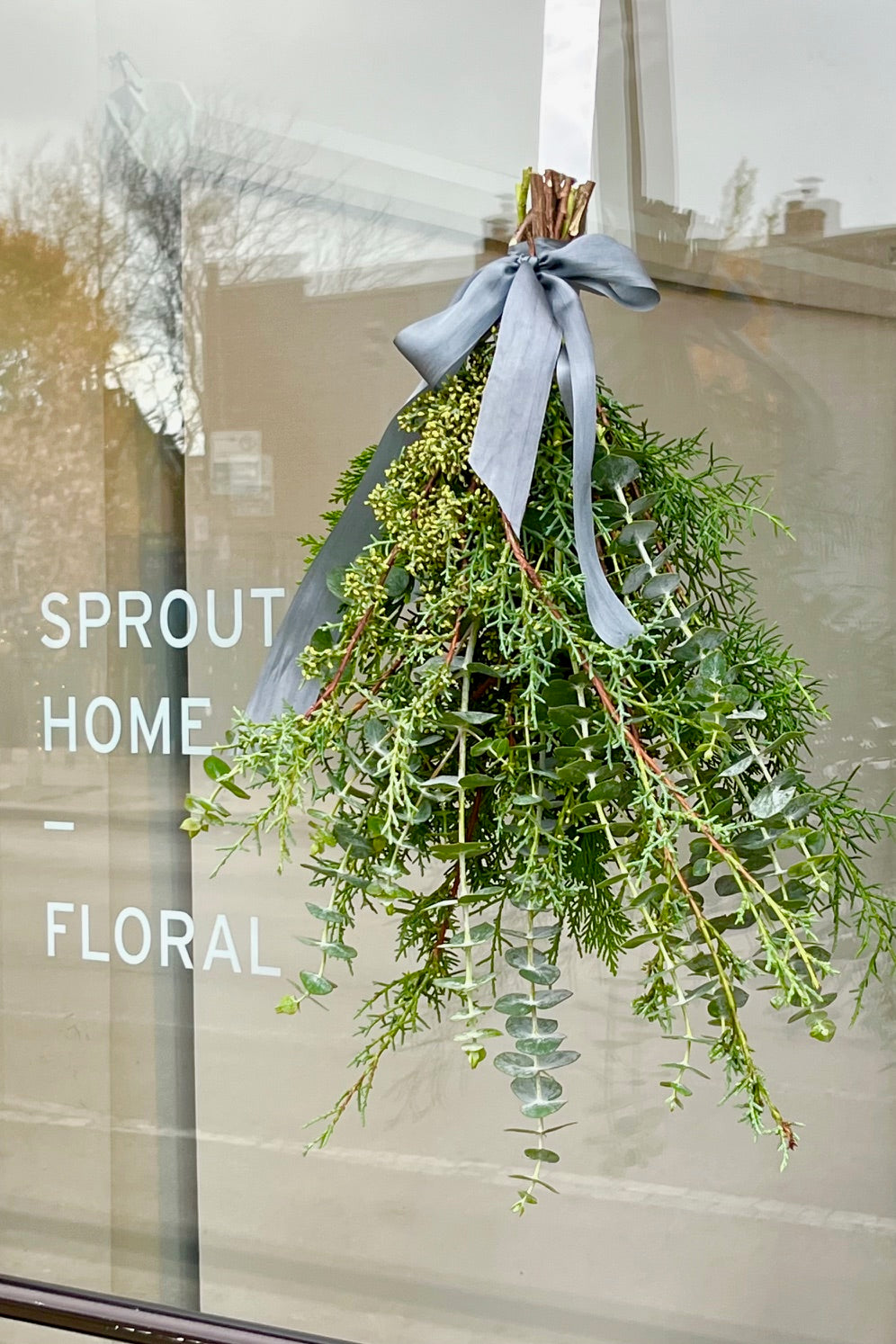 Hand holding a mixed greenery bouquet with eucalyptus, cypress, cedar tied with ribbon hanging on the glass door to the Sprout Home Floral store. ©Sprout Home #size_$35