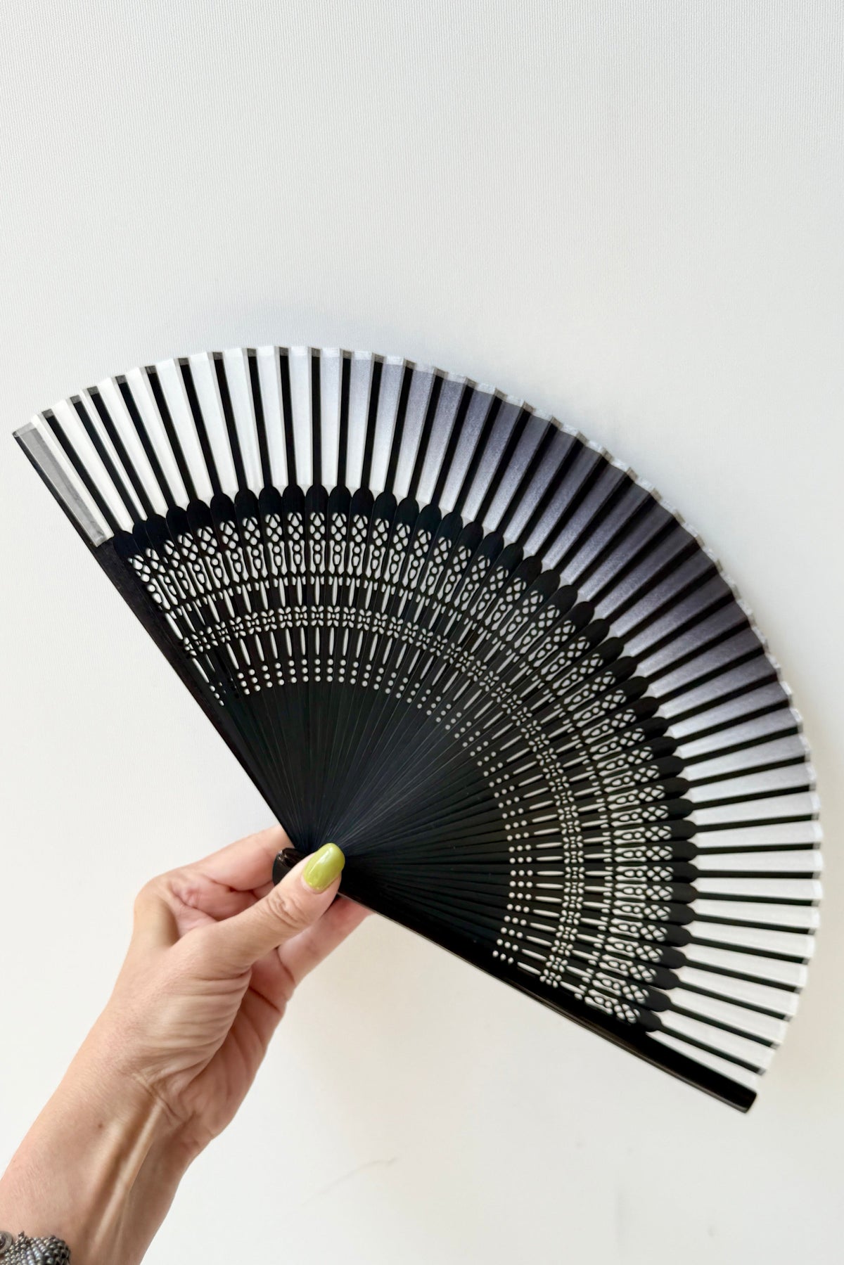 A hand holding a Silk Folding Fan White Silk w/ Black Bamboo with intricate white geometric patterns on the underside, set against a plain white background. ©Sprout Home