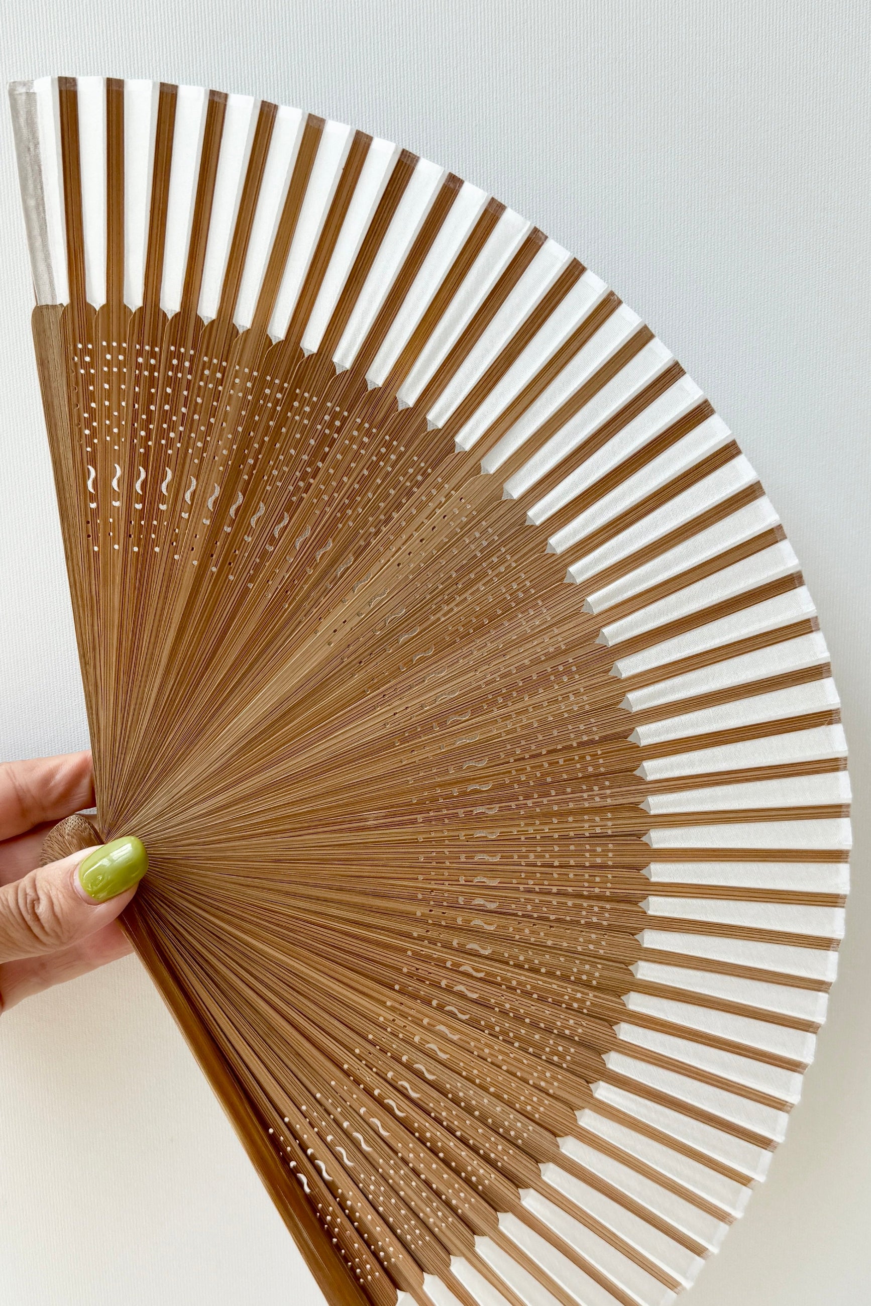 A traditional Silk Folding Fan Light Beige w/ Brown Bamboo made of light brown wood, held open by a hand with green painted fingernails against a plain white background. ©Sprout Home