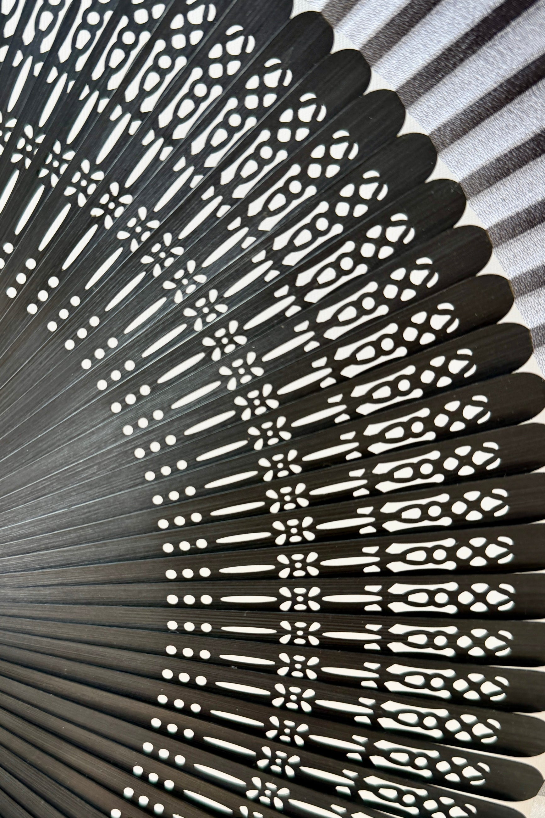 Close-up of a Silk Folding Fan White Silk w/ Black Bamboo with intricate white cutout patterns on the ribs, arranged in a radial fan shape against a light gray background. ©Sprout Home