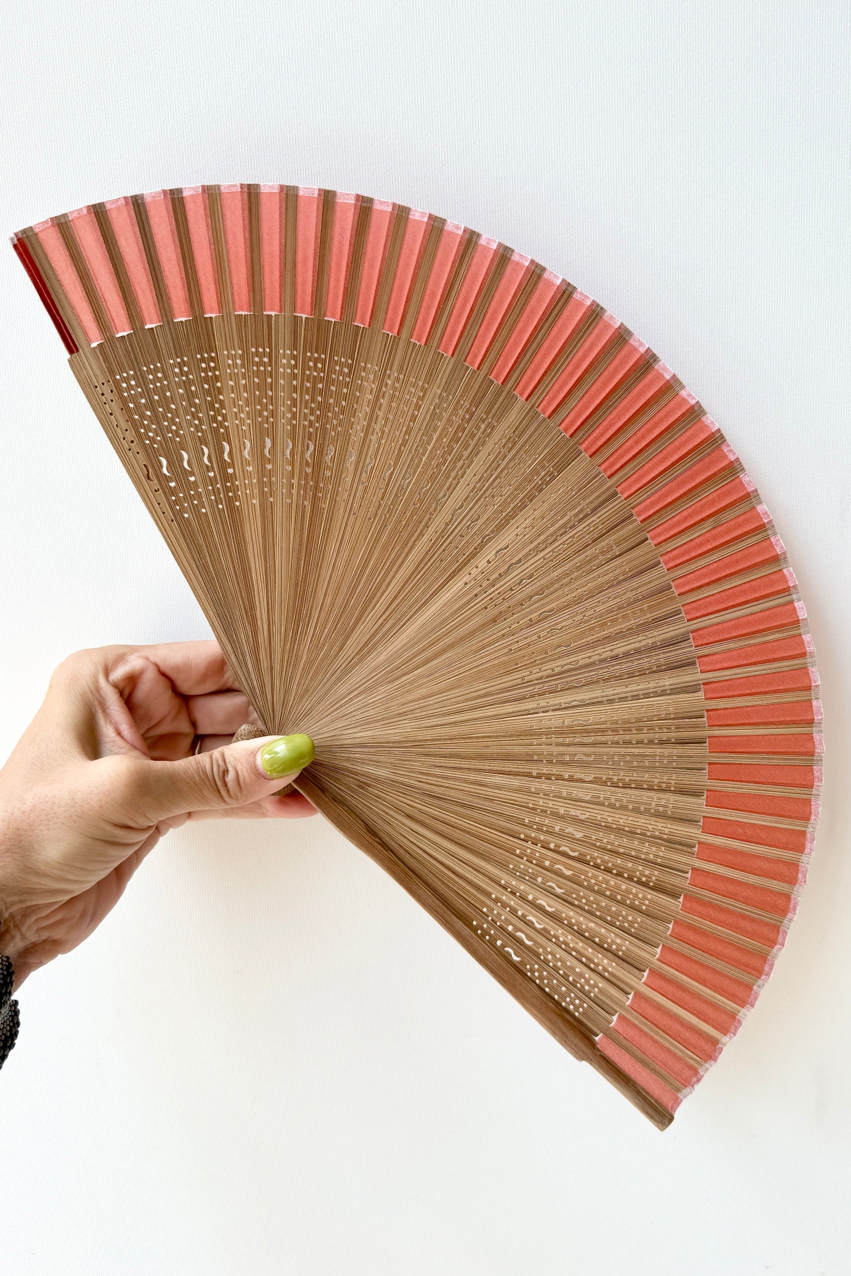 A hand with lime green nail polish holds a Silk Folding Fan Harvest Brown w/ Brown Bamboo with light brown wooden ribs and coral-pink paper blades against a plain white background. ©Sprout Home
