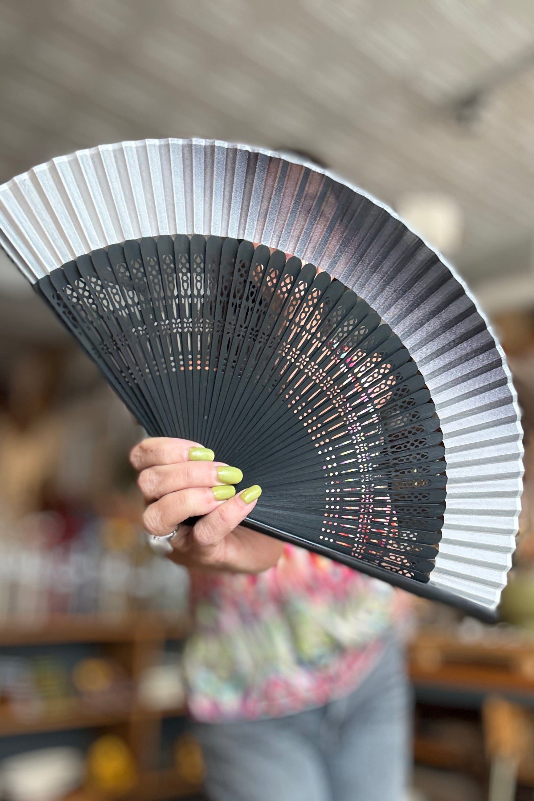 A hand holding a Silk Folding Fan White Silk w/ Black Bamboo with a black and white printed design, set against a blurred indoor background. ©Sprout Home