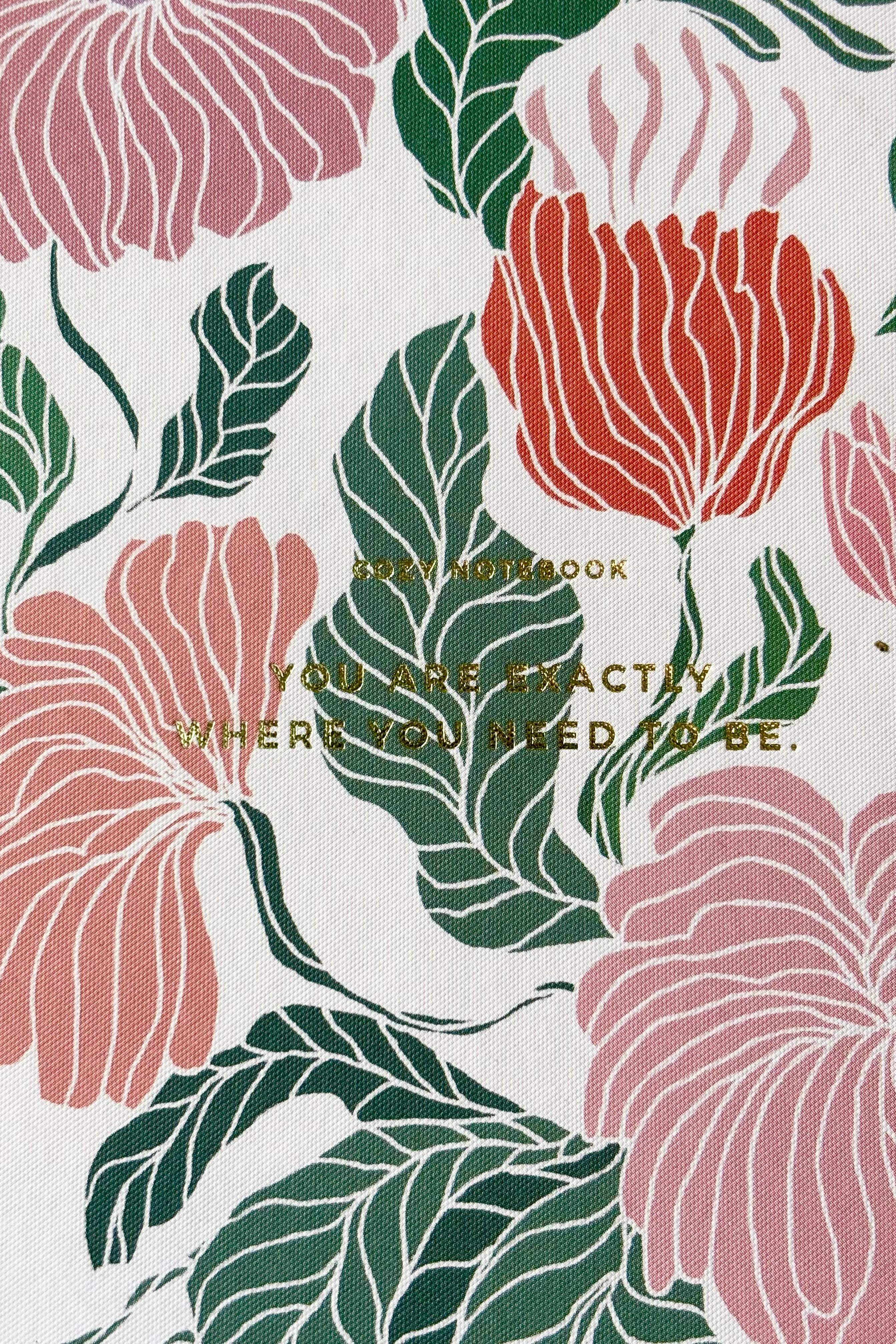 Close up of hard cover notebook with pink, red and green floral motif against a pink background with the phrase "You are exactly where you need to be" in gold lettering