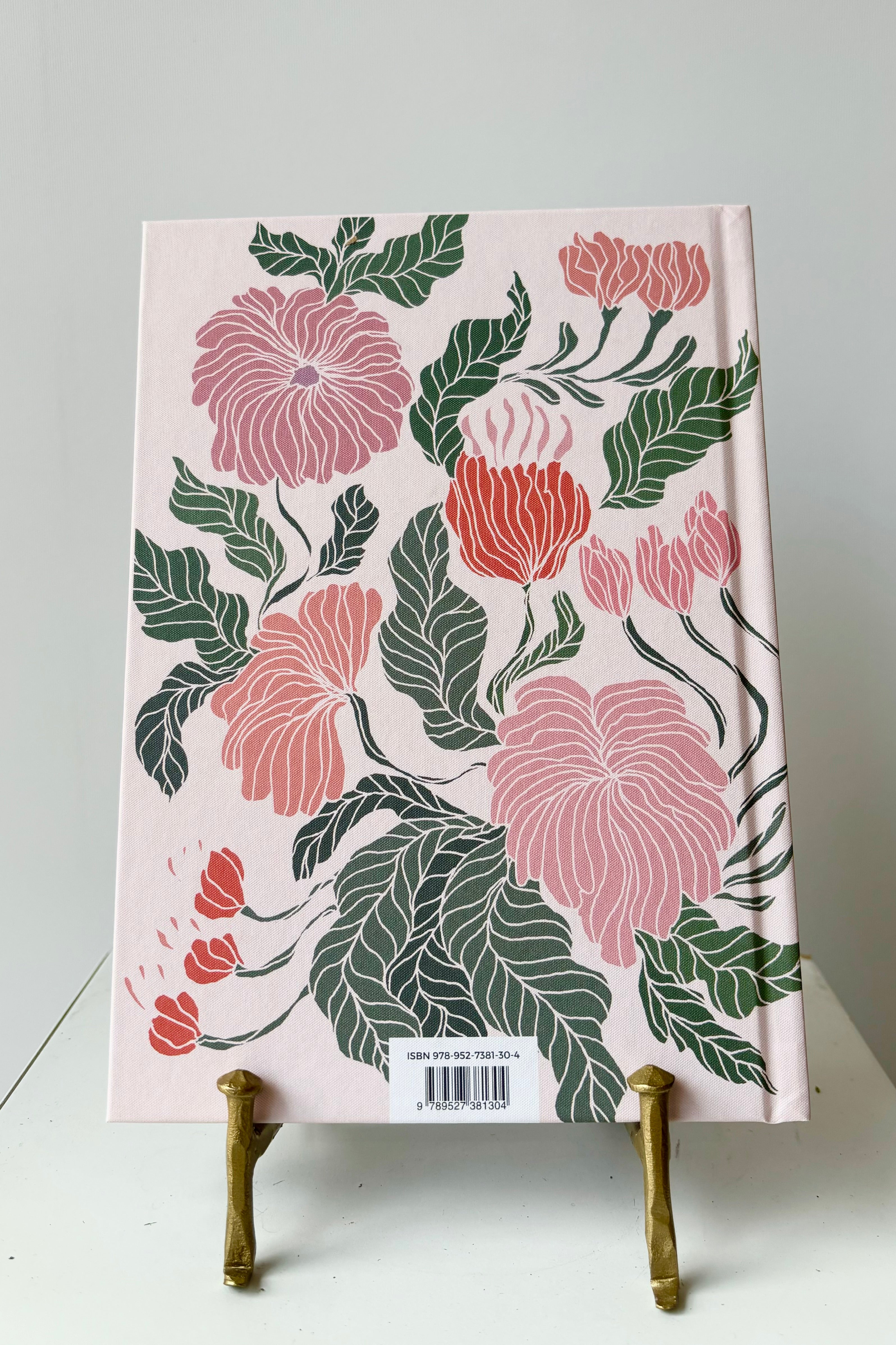 Back cover of notebook with pink, red and green floral motif against a pink background with the phrase "You are exactly where you need to be" in gold lettering, displayed in a gold bookstand against white background