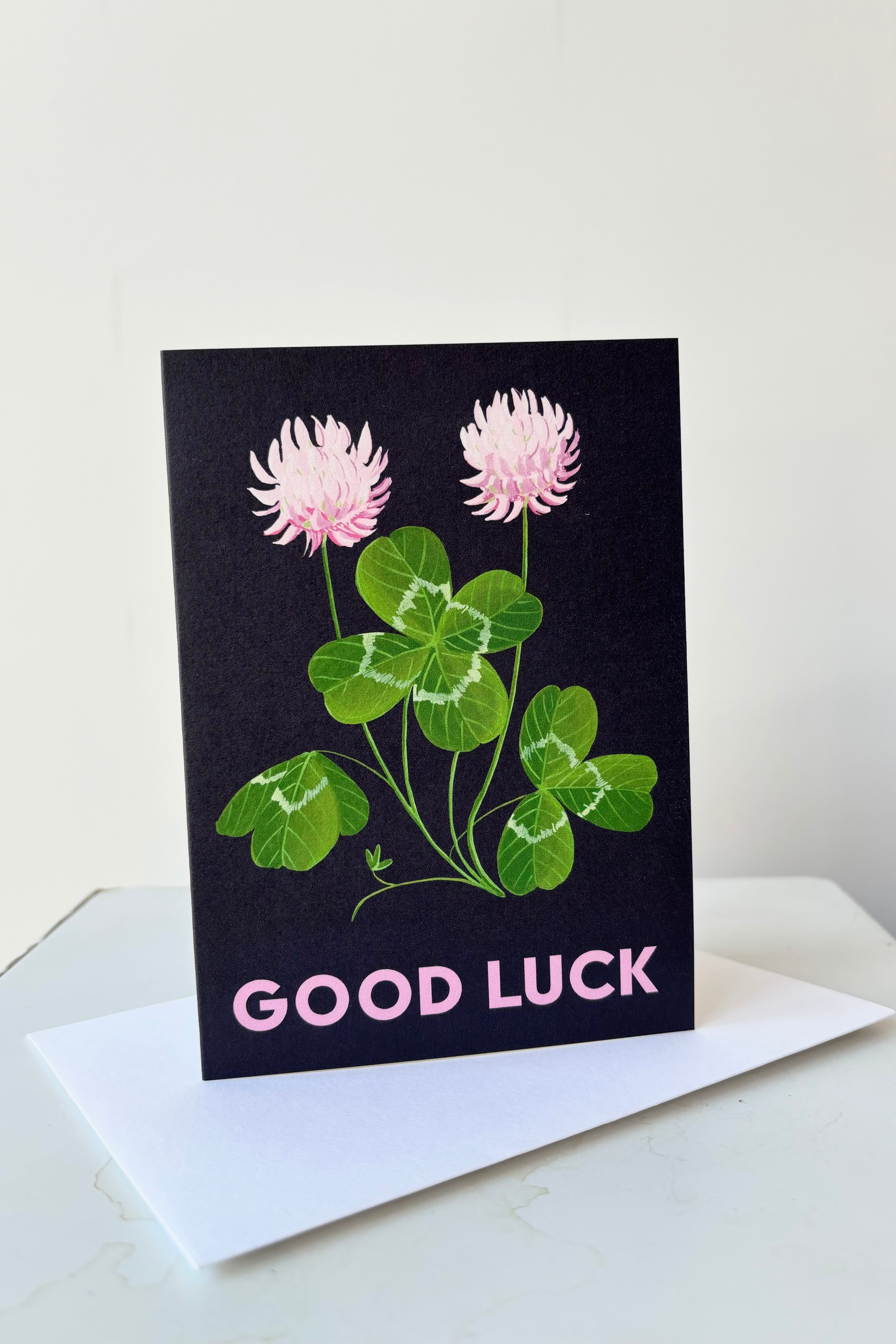 Clover greeting card with image of clover plant with pink flowers on dark blue background with the text Good Luck on the front, blank inside against white background displayed on top of white envelope.