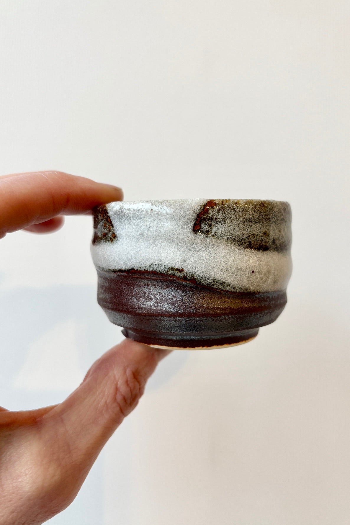 Hand golding the Bizan sake cup with clay body and brown, white and gray glazing against white background. ©Sprout Home