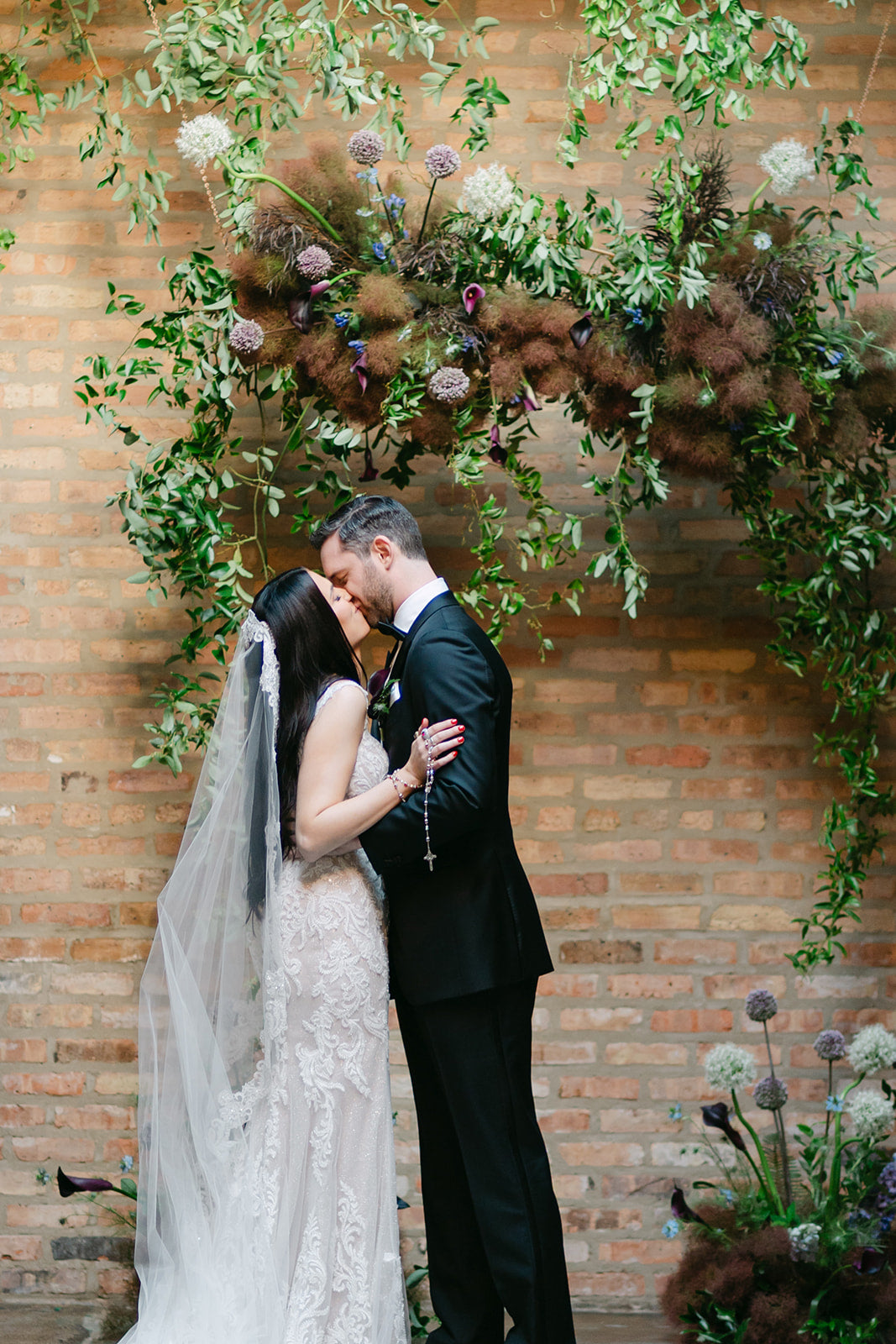 Couple kissing under a floral archway designed by Sprout Home with a brick wall background at The Arbory, a wedding venue in Chicago ©Devae Photography