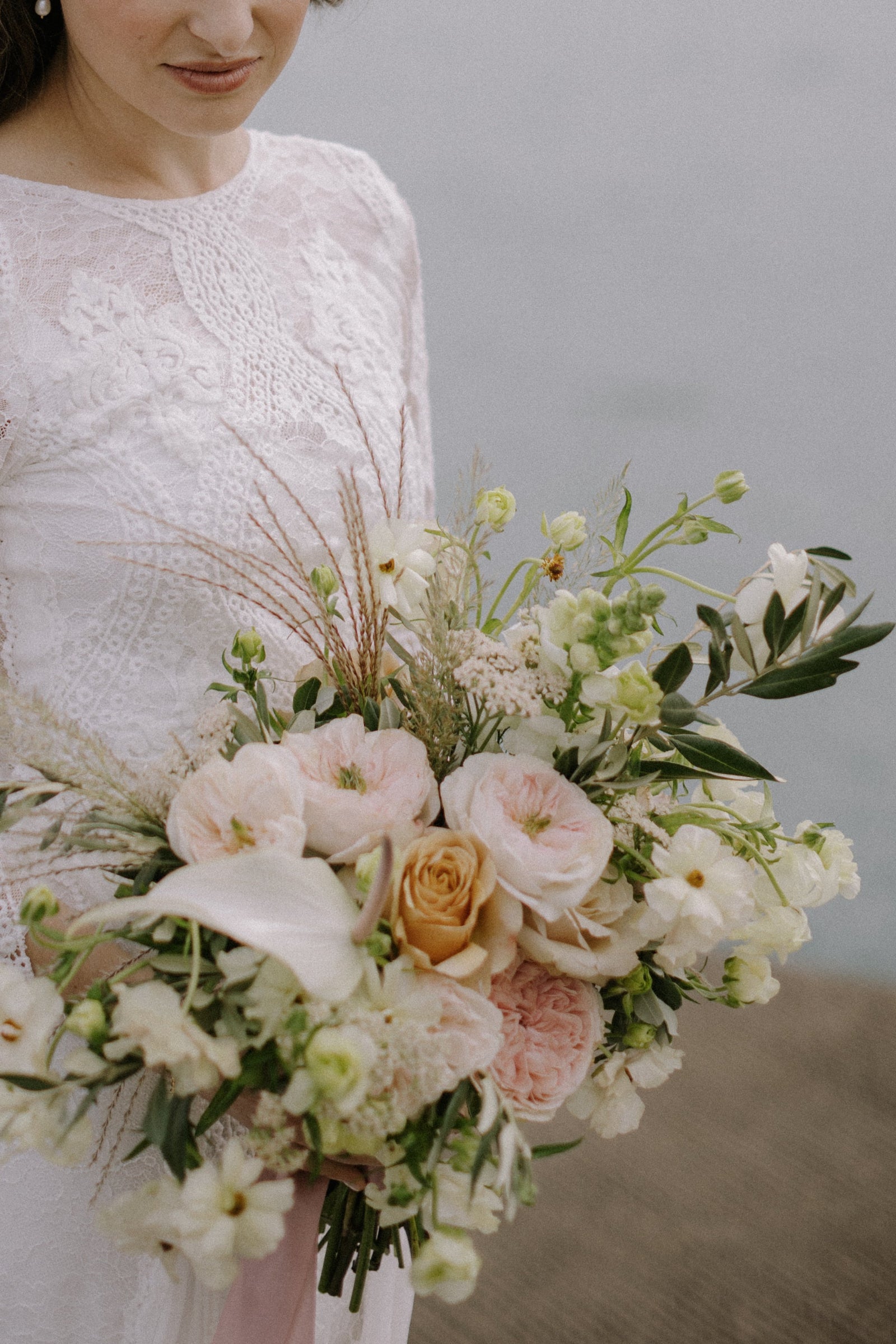 Kroster wedding with the bride in lace, holding a white and light pastel floral with hints of grass plumes. arrangement by Sprout Home . Photograph by Samantha Elizabeth Creative