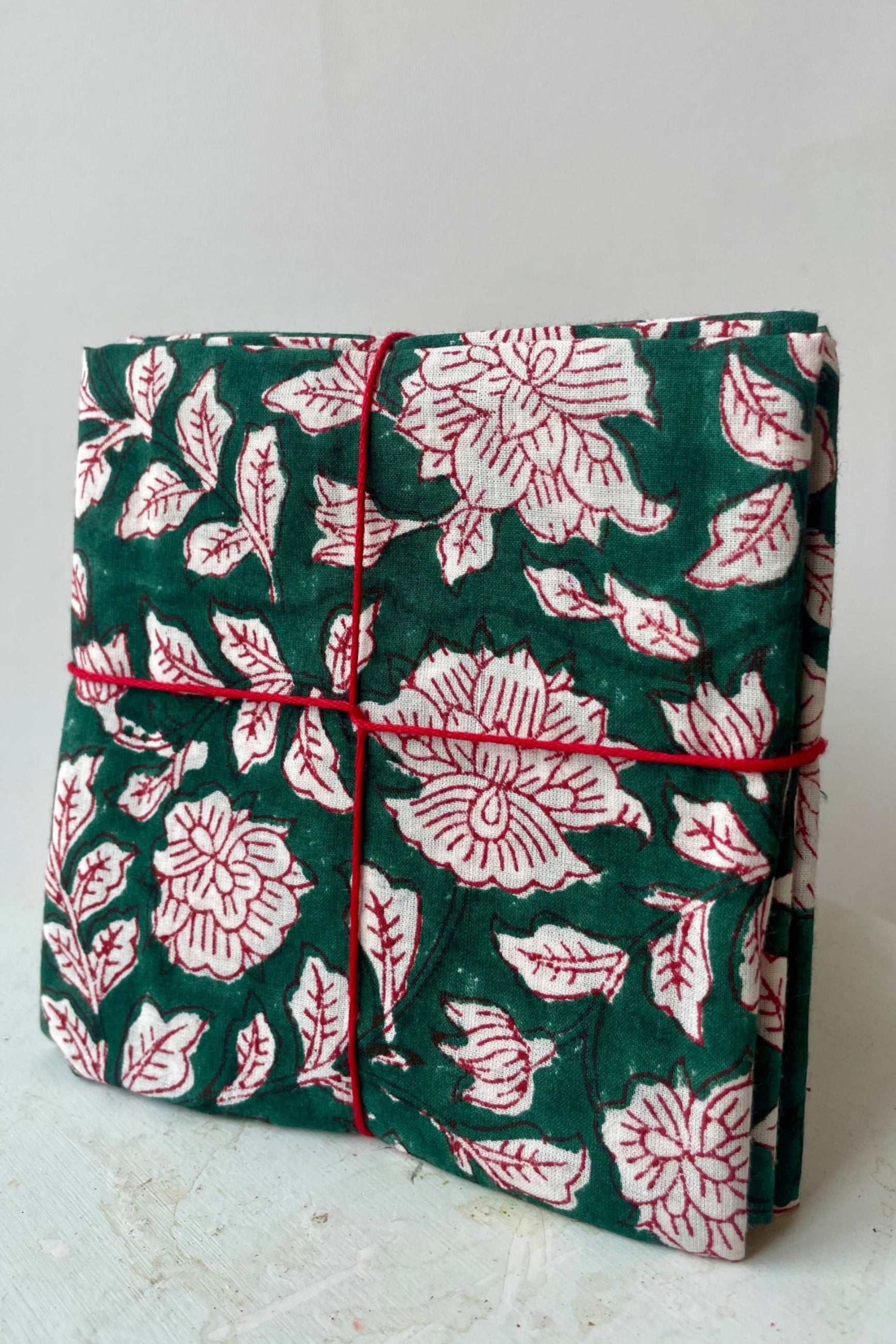 Photo of a set of cloth napkins folded as a square. A floral pattern of red and white on a green background. Colorful floral napkins are shown in front of a white wall. ©Sprout Home