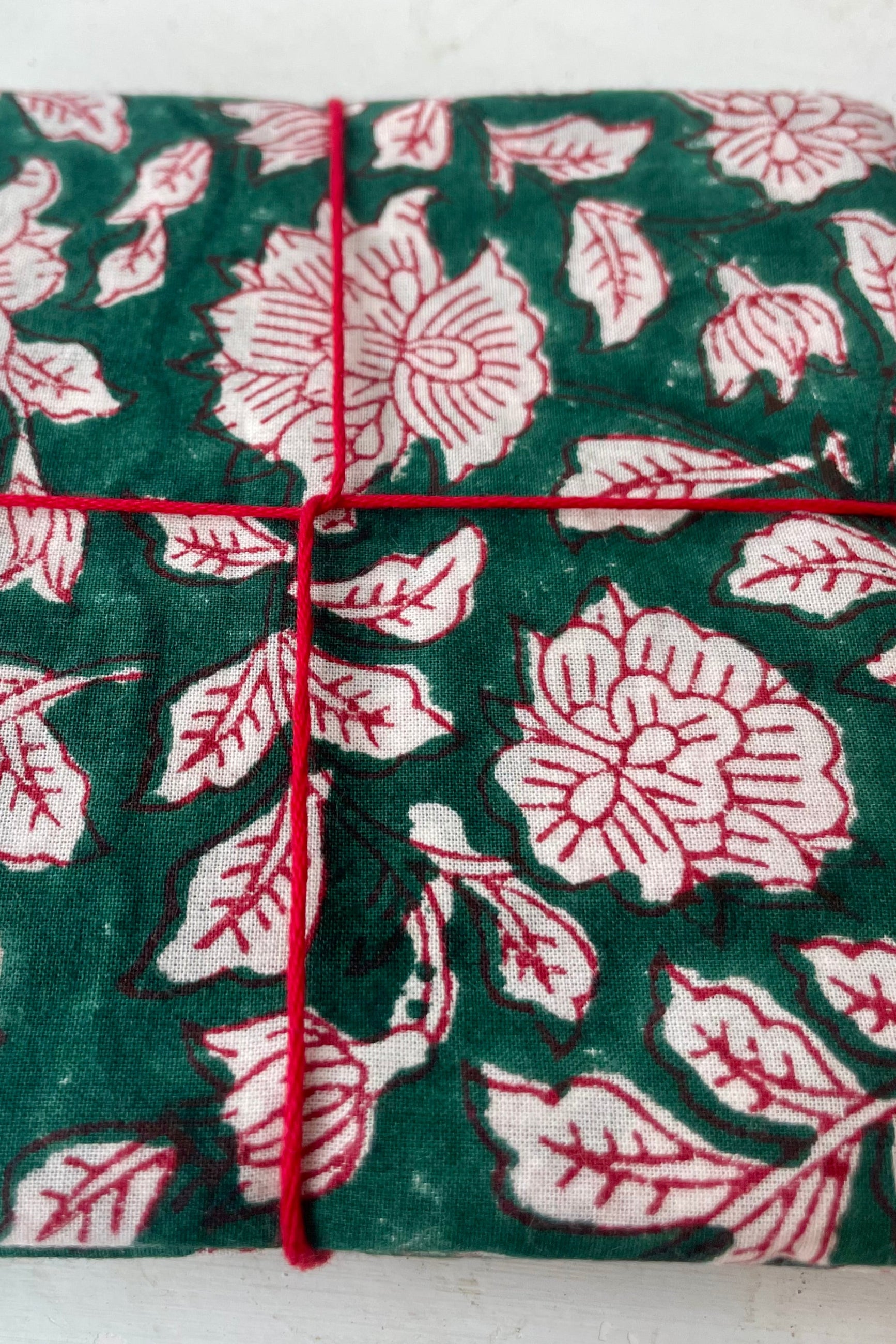 Close Photo of a set of cloth napkins folded as a square. A floral pattern of red and white on a green background. Colorful floral napkins are shown in front of a white wall. ©Sprout Home
