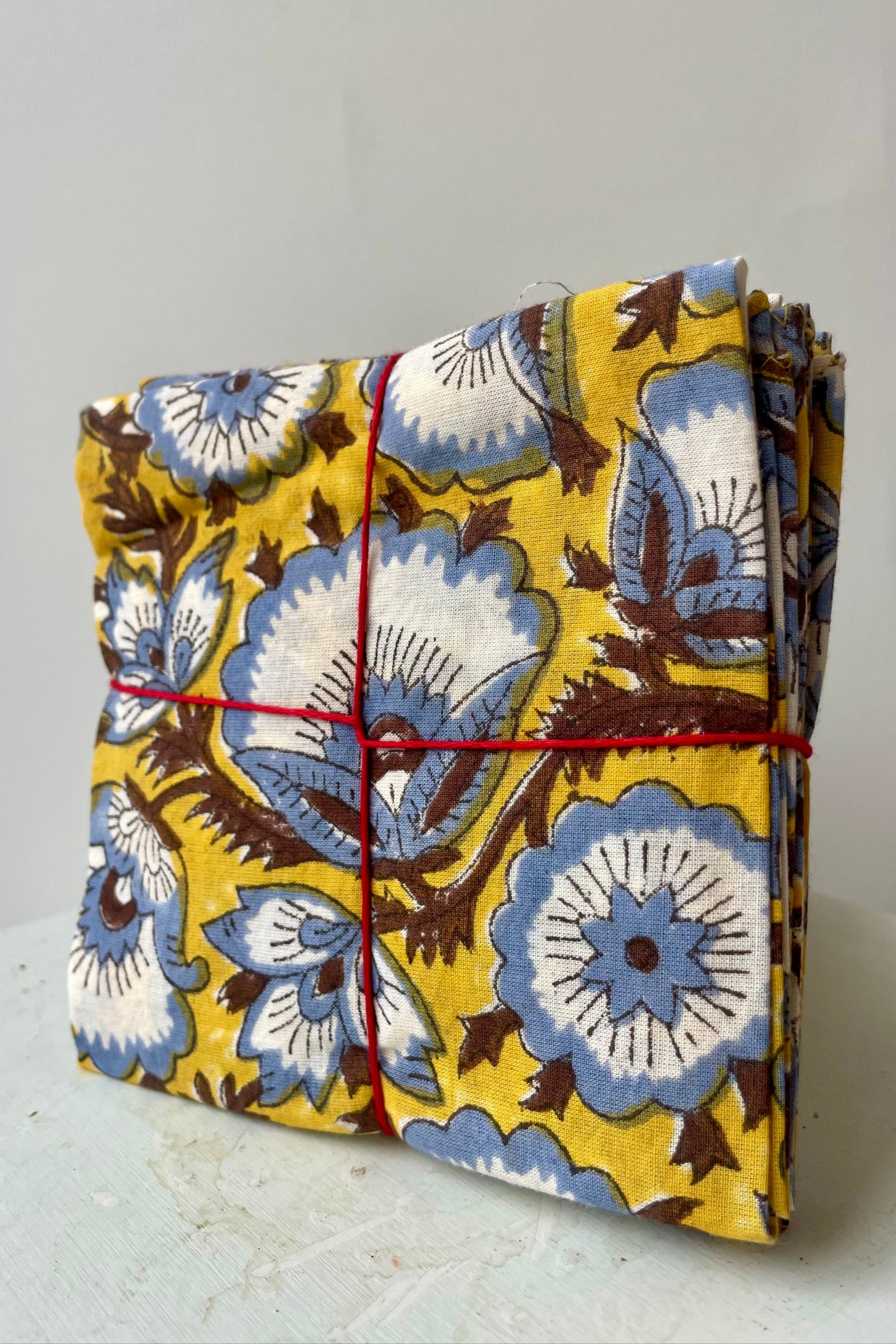 Photo of a set of cloth napkins folded as a square. A floral pattern of blue and brown on a yellow background. Colorful floral napkins are shown in front of a white wall. ©Sprout Home