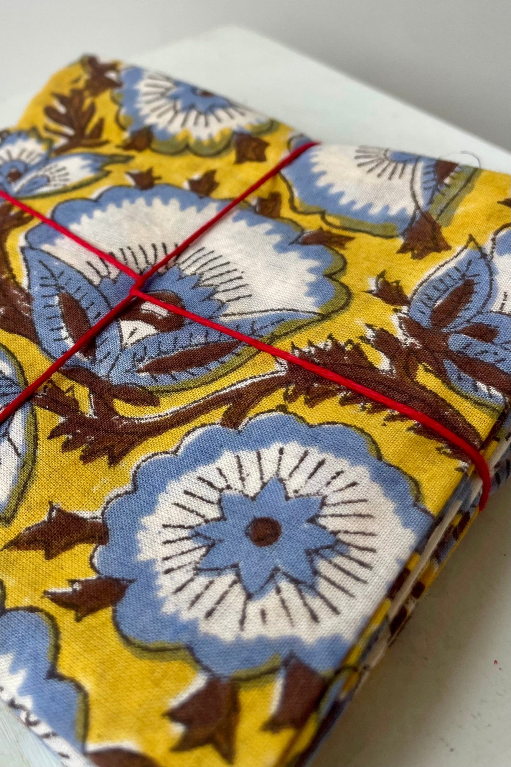 Close Photo of a set of cloth napkins folded as a square. A floral pattern of blue and brown on a yellow background. Colorful floral napkins are shown in front of a white wall. ©Sprout Home