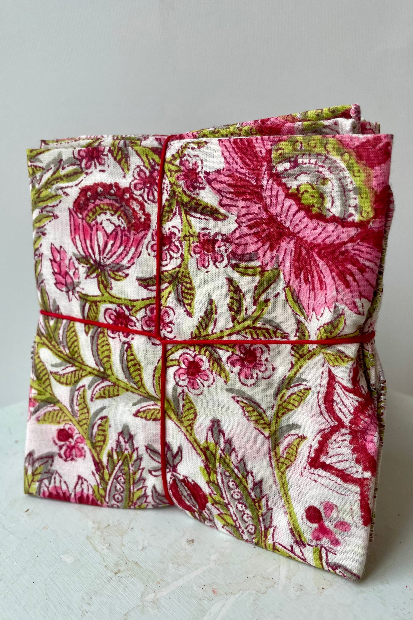 Photo of a set of cloth napkins folded as a square. A floral pattern of pink and green on a white background. Colorful floral napkins are shown in front of a white wall. ©Sprout Home