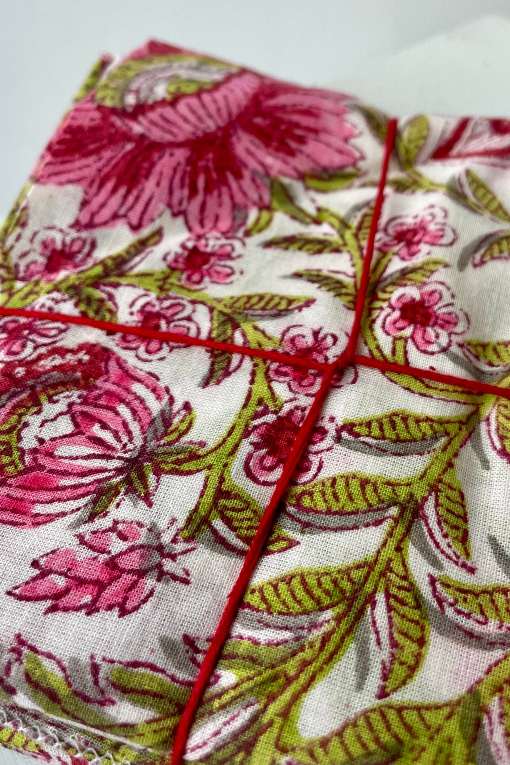 Close Photo of a set of cloth napkins folded as a square. A floral pattern of pink and green on a white background. Colorful floral napkins are shown in front of a white wall. ©Sprout Home
