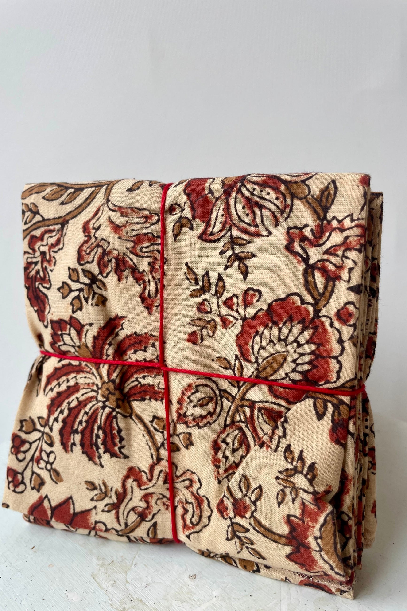 Photo of a set of cloth napkins folded as a square. A floral pattern of sienna and brown on a tan background. Colorful floral napkins are shown in front of a white wall. ©Sprout Home