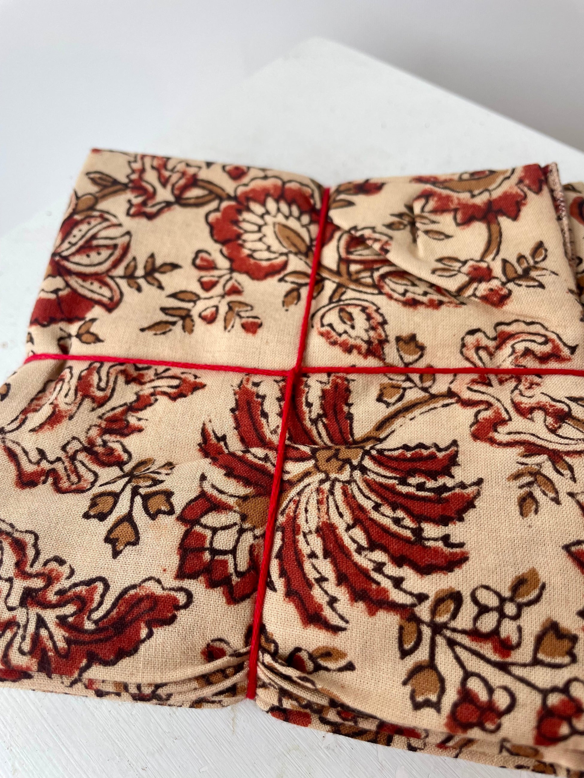 Close Photo of a set of cloth napkins folded as a square. A floral pattern of sienna and brown on a tan background. Colorful floral napkins are shown in front of a white wall. ©Sprout Home