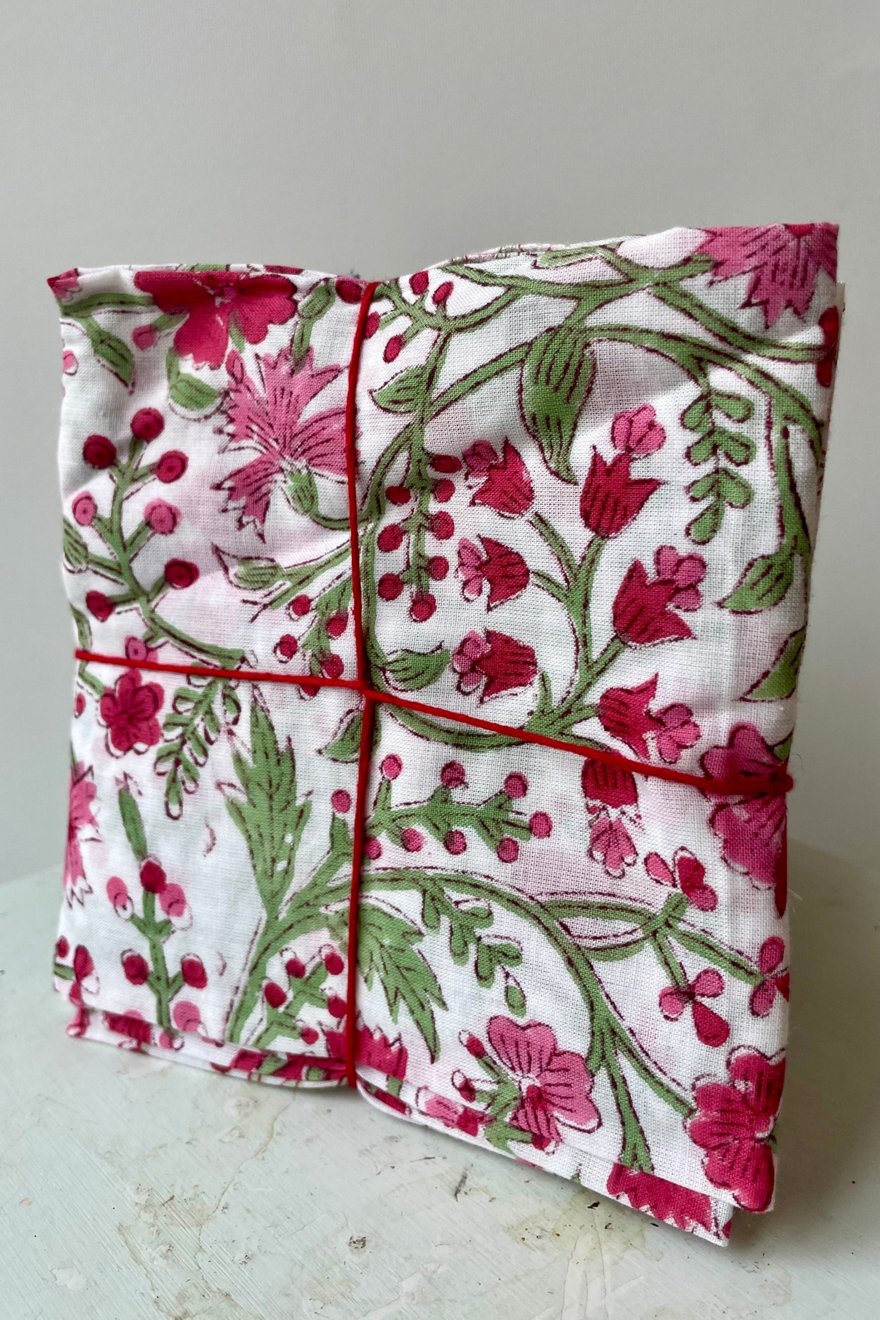 Photo of a set of cloth napkins folded as a square. A floral pattern of pink and green on a white background. Colorful floral napkins are shown in front of a white wall. ©Sprout Home