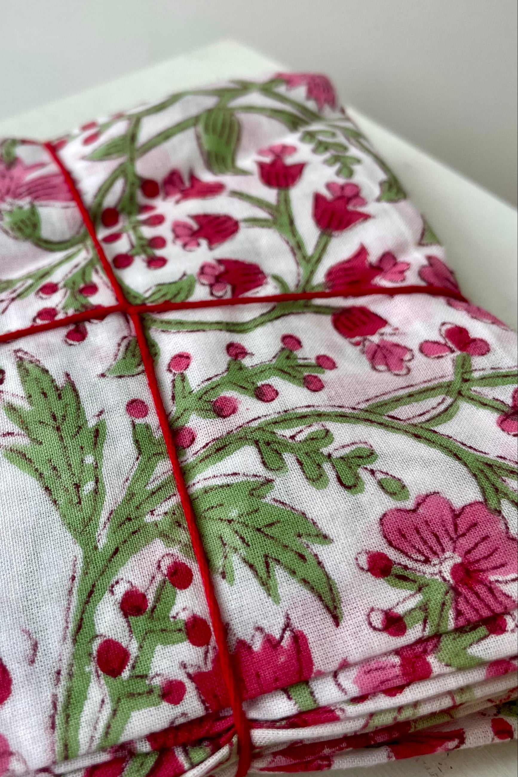 Close Photo of a set of cloth napkins folded as a square. A floral pattern of pink and green on a white background. Colorful floral napkins are shown in front of a white wall. ©Sprout Home