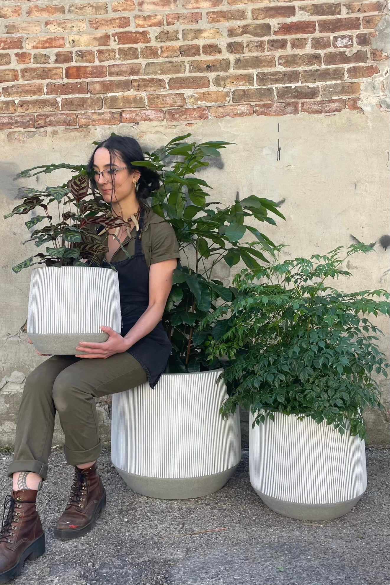 A person sitting with three sizes of white and gray striped Harith pots and plants against a cement and brick weall.