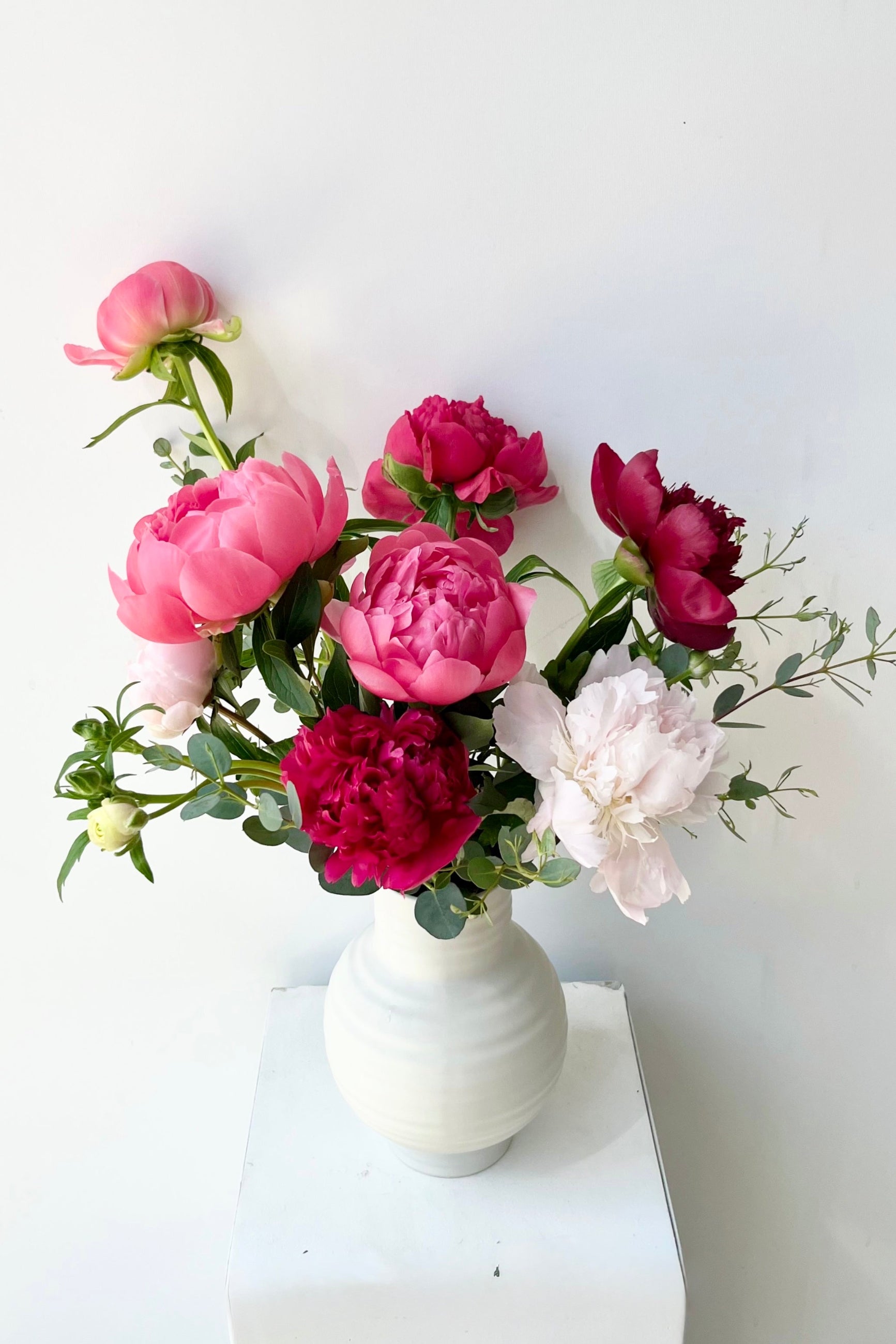 Bouquet of pink, red, and white Paeonia flowers in a white vase on a white surface with a light gray background ©Sprout Home #size_$100