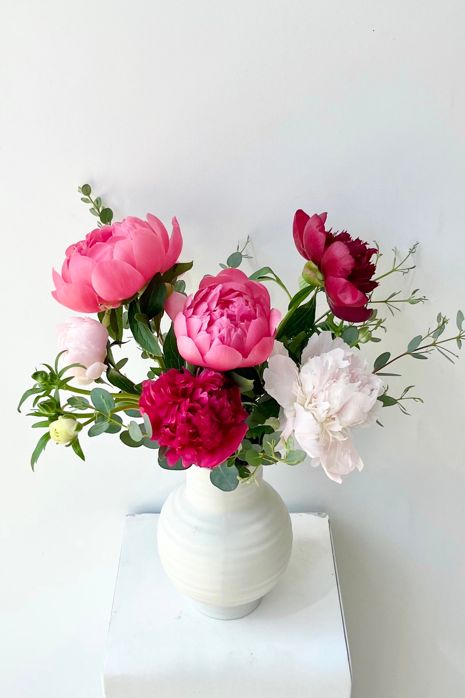 Bouquet of pink, red, and white Paeonia flowers in a white vase on a white surface with a light gray background ©Sprout Home #size_$75