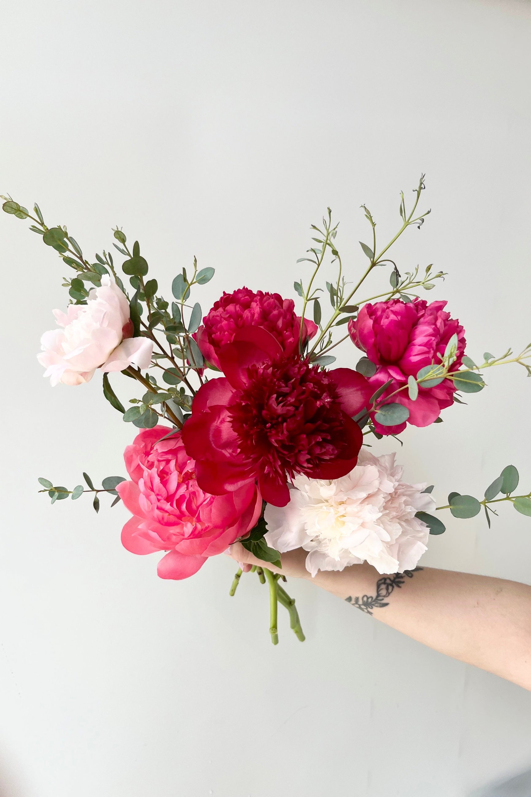 Hand holding a bouquet of pink, red, and white Peony flowers against a light gray background ©Sprout Home #size_$75