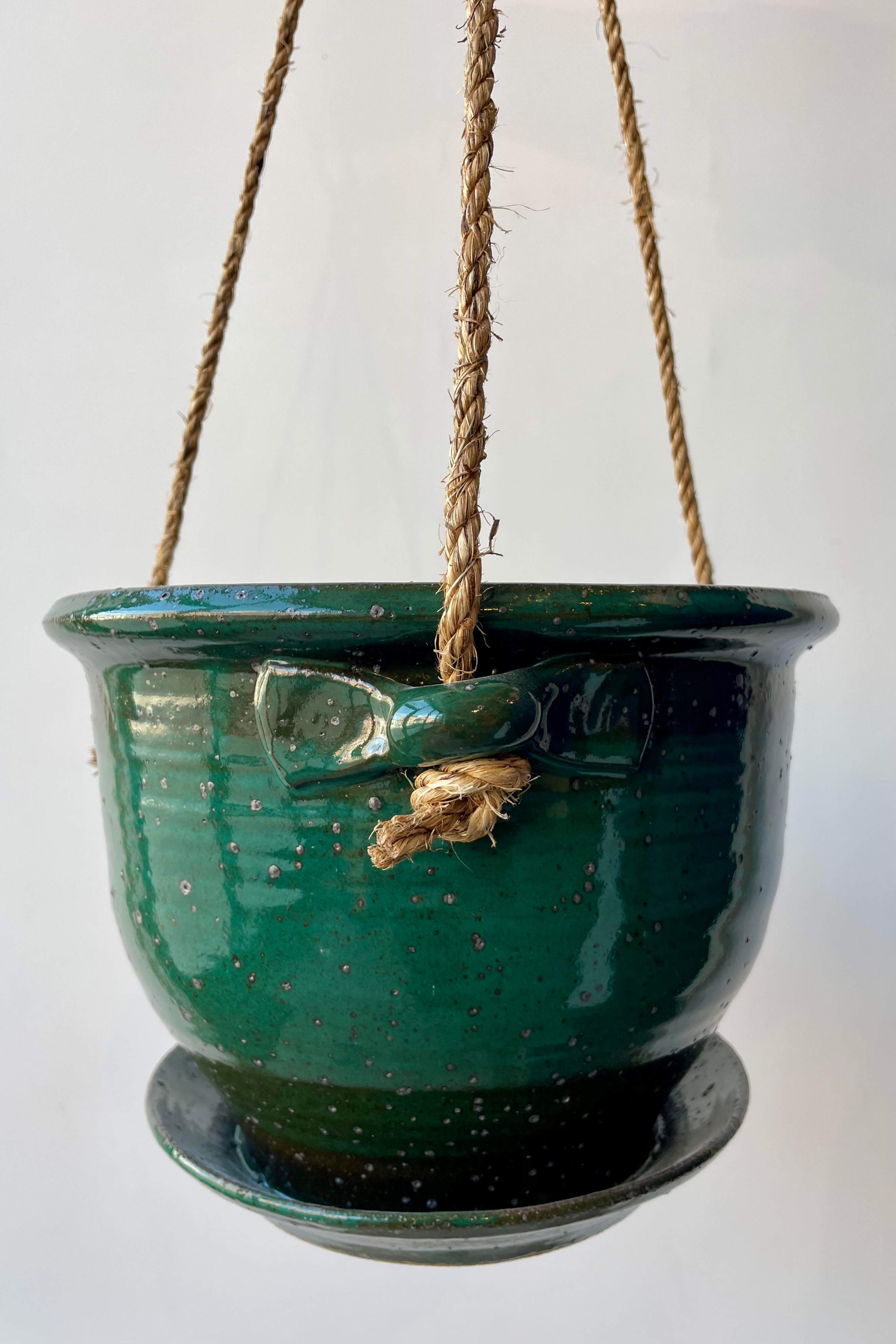 Eight inch clay planter with drainage hole and attached tray in a jade green glaze flecked with silver specks against a white background. The planter has a jute cord attached at three points to hang.