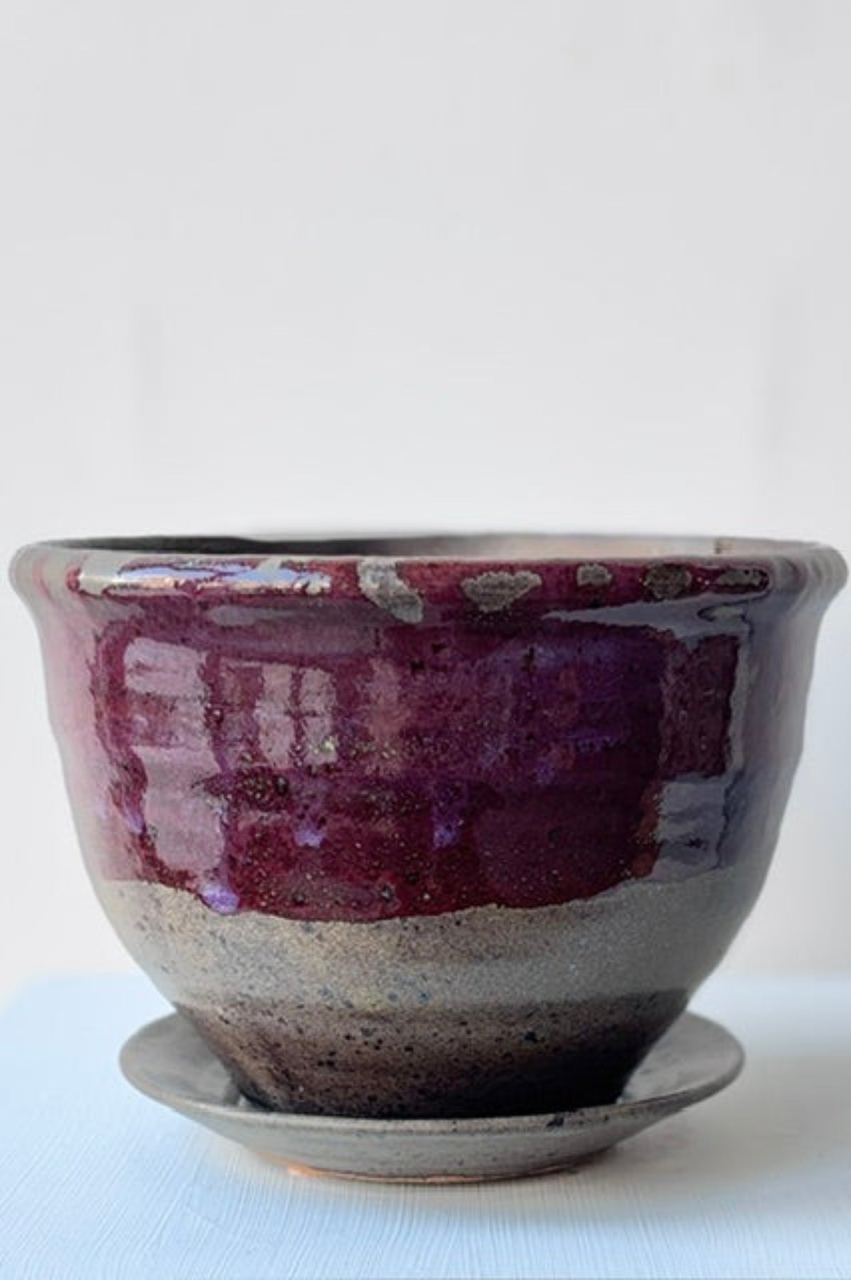 A handmade ceramic planter pot with a matching saucer, featuring a glossy deep purple and dark brown glaze over a speckled gray clay base, set against a plain white background ©Sprout Home