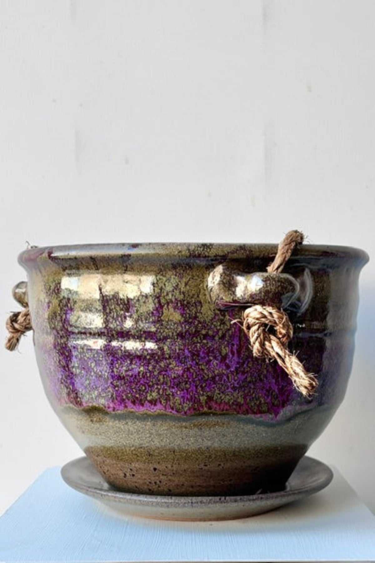 A handmade ceramic planter pot with a matching saucer, featuring a metallic bronze and purple glaze with two decorative rope handles, set against a plain white background.