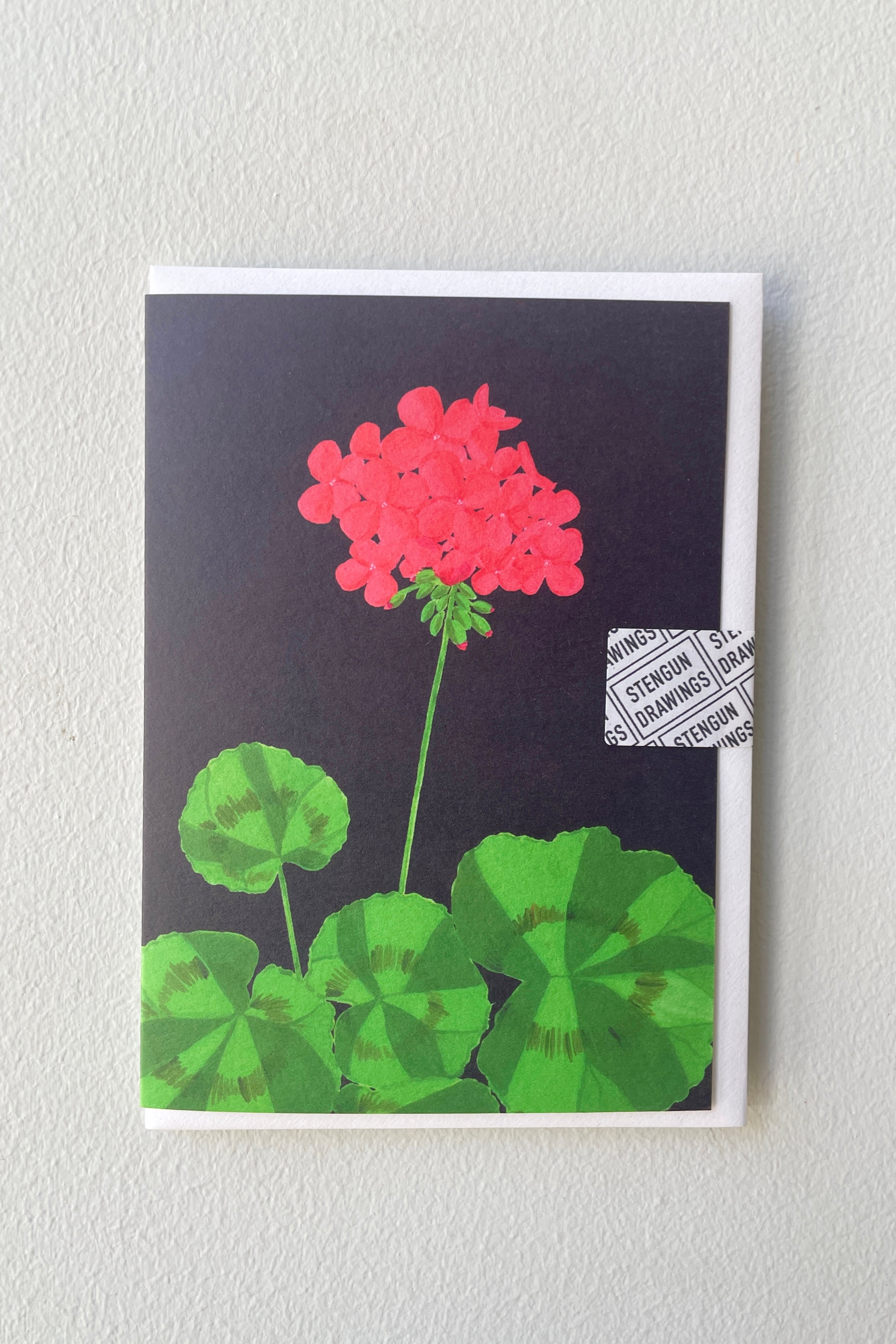 The Stengun Barbican Geranium greeting card against a white wall.