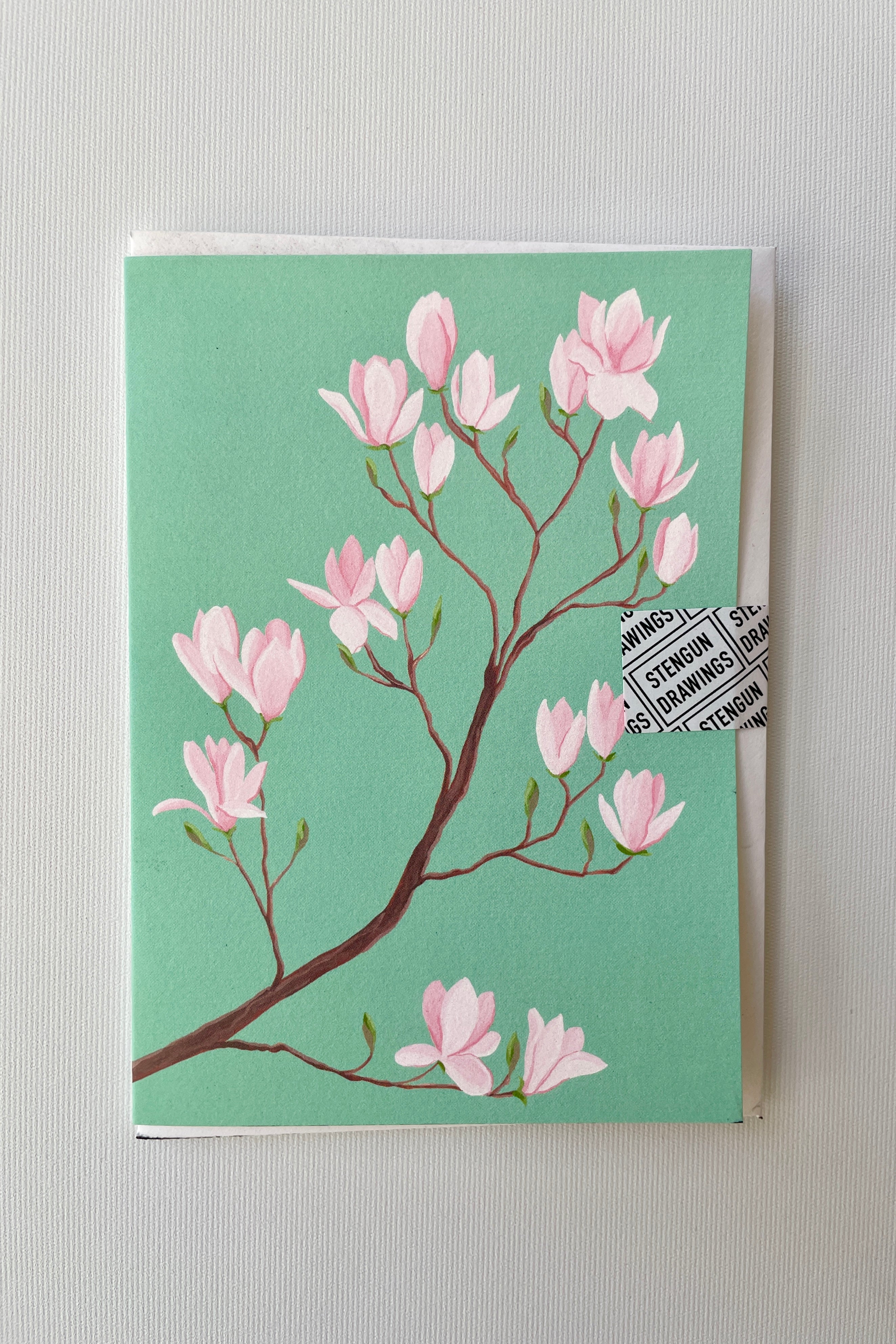 A Magnolia greeting card by Stengun at Sprout Home with its turquoise background and pink flowers.