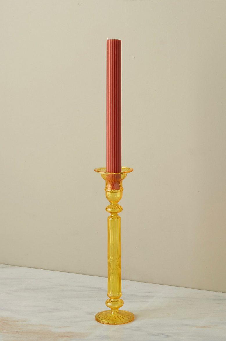 A Nahlia Daffodil colored glass candlestick holder with a terracotta colored taper candle unlit.