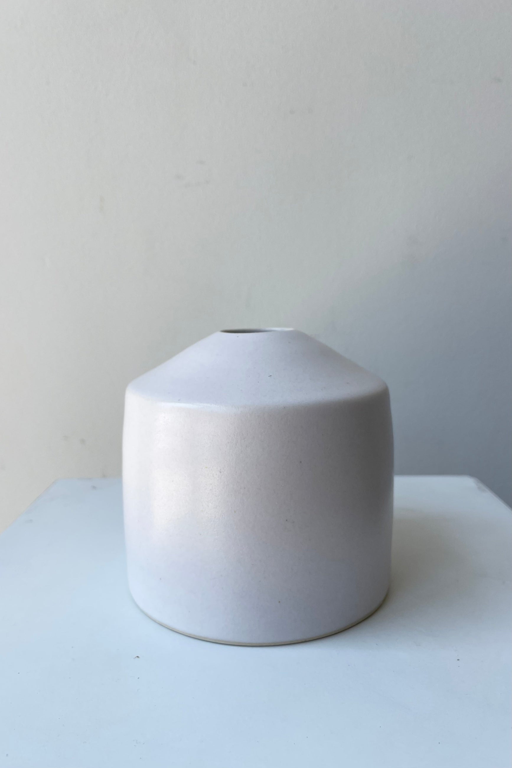One small ceramic vase sits on a white surface in a white room. The vase is cylindrical and tapers off at the top to a narrow opening. The vase is white with a small ring of unglazed clay at the bottom. It is photographed straight on. ©Sprout Home