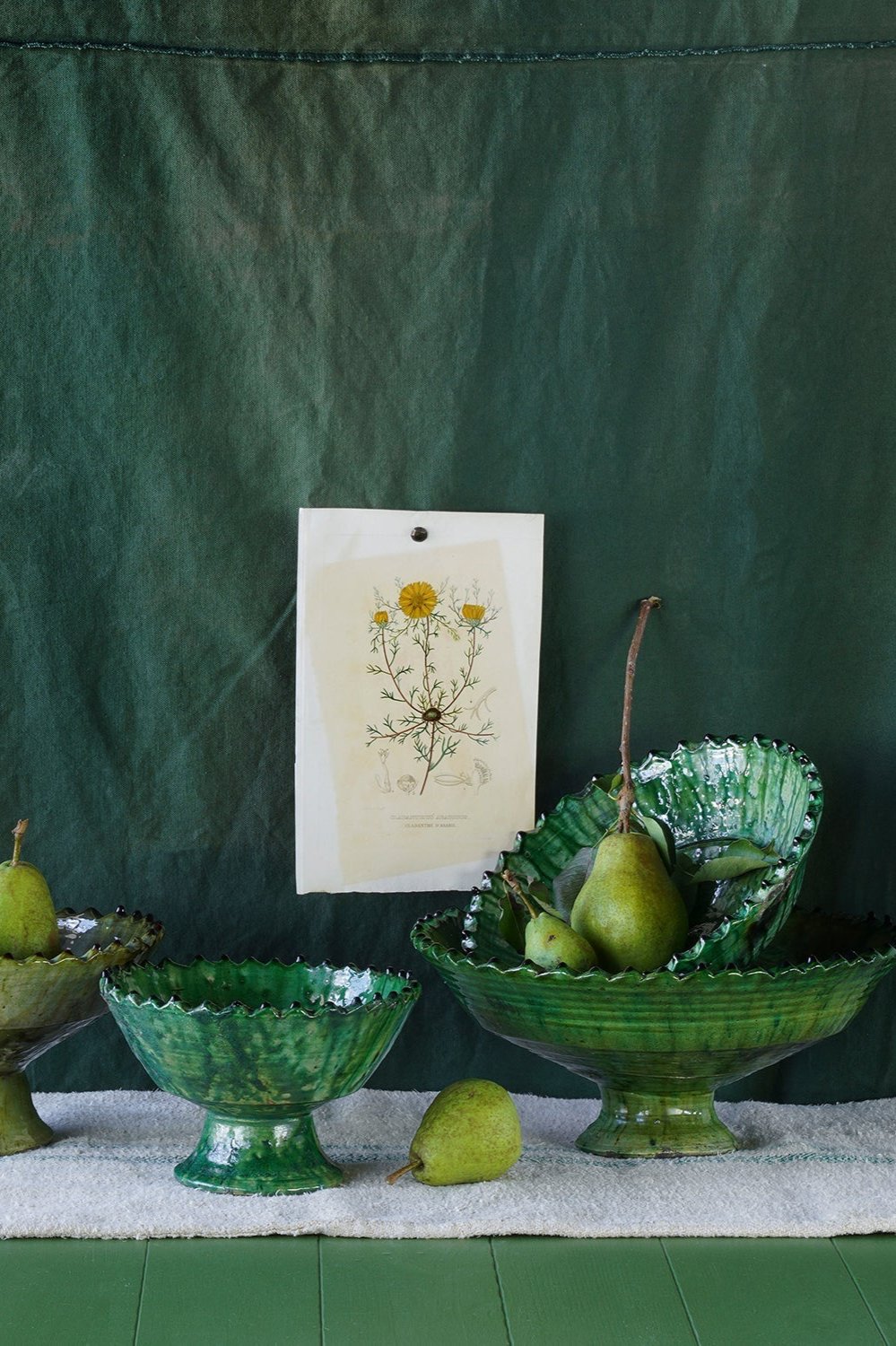 Various sized green tamegroute compote bowls against a green fabric background, a print with dandelions and pears. ©Blue Ocean Traders