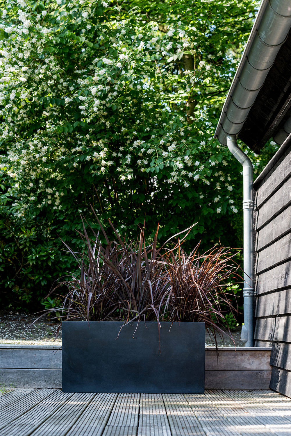 Jort Jumbo Black Large planter with decorative grasses on a wooden deck against a backdrop of greenery and a building. ©Pottery Pots