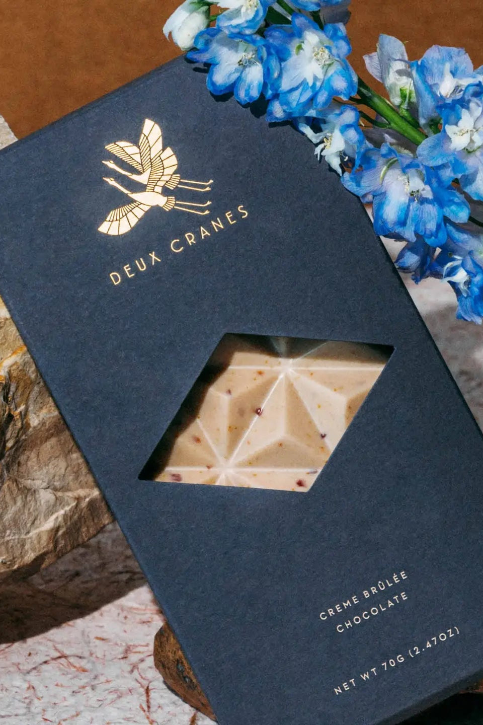 Photo of a Deux Cranes Creme Brulee Chocolate bar in a dark blue box with blue Delphinium flower in front of a stone background.