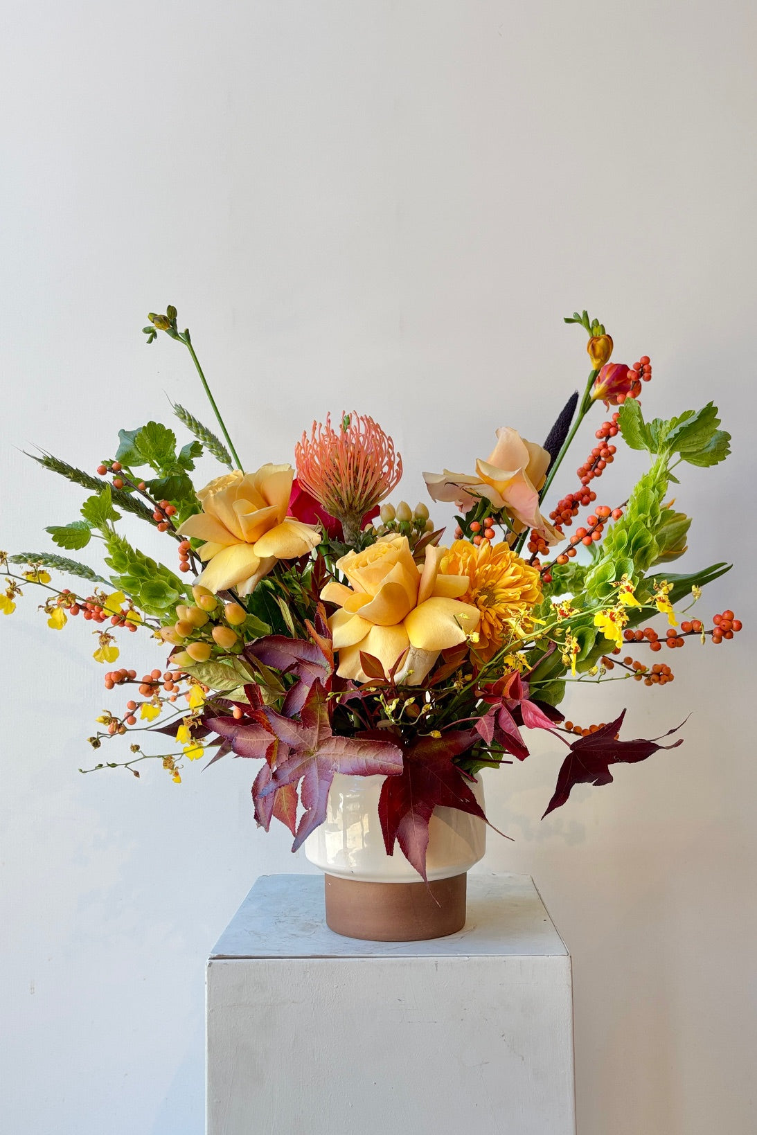 Colorful custom floral arrangement by Sprout Home featuring fall burgundy foliage and warm golden roses in a white vase and brick colored vase on a light off-white background ©Sprout Home #size_$160