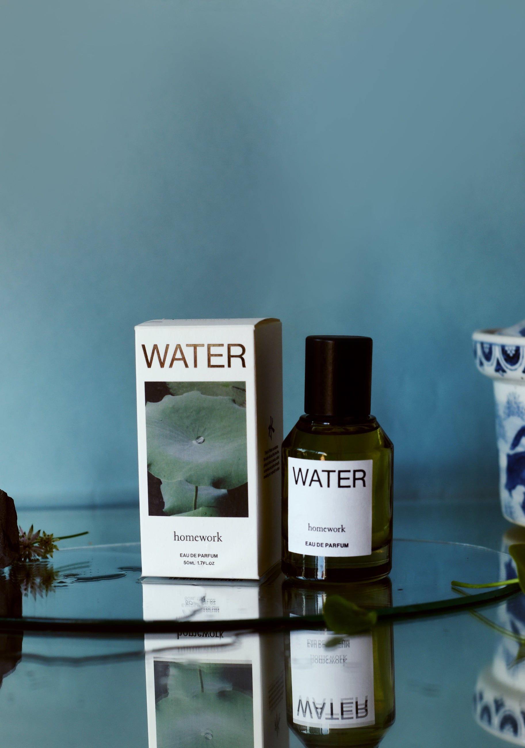 Homework Water Perfume bottle and packaging side by side sitting on a mirror in a blue themed setting ©Homework