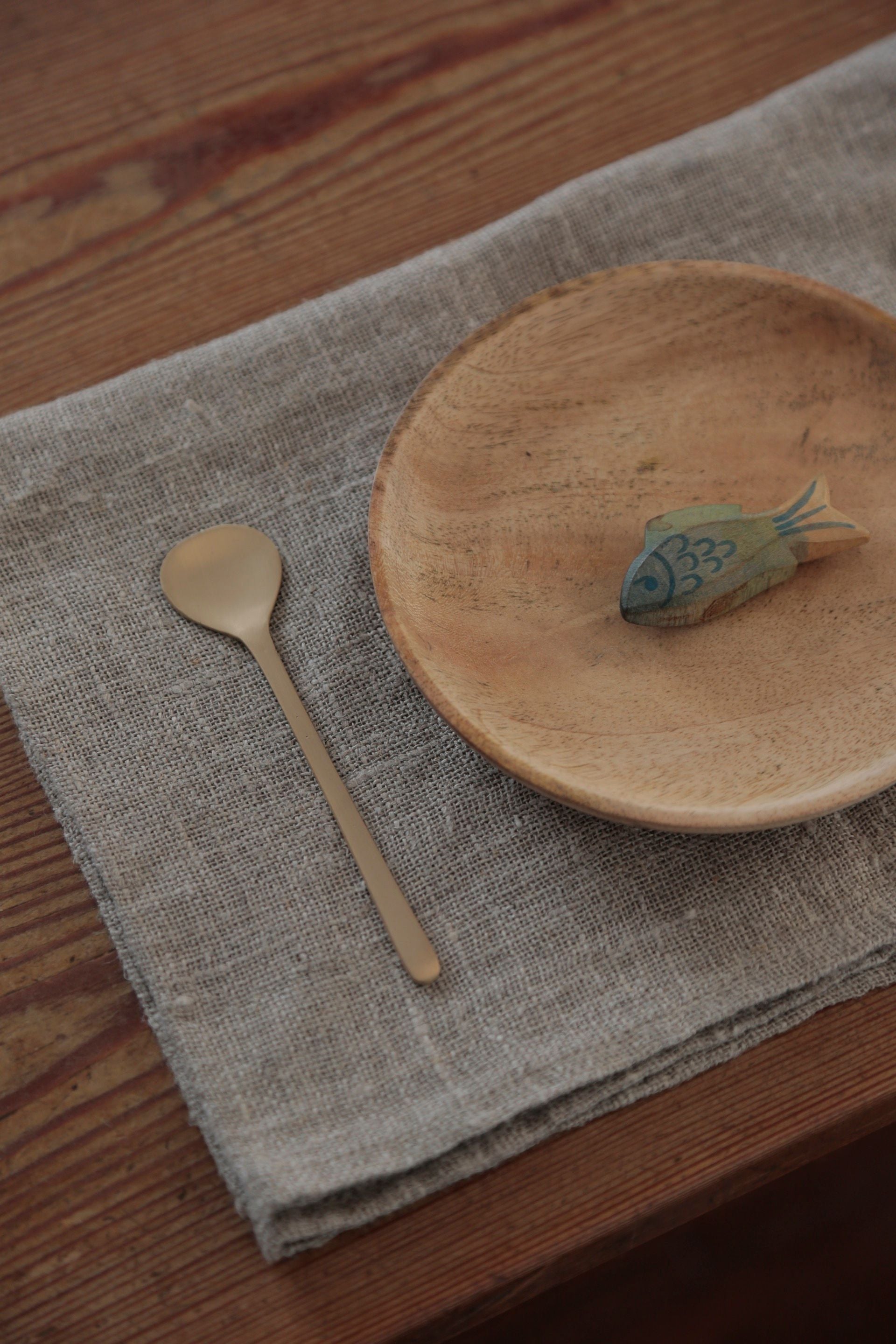 Brass dessert spoon by Fog Linen Work sitting on a linen cloth and next to a wood plate with a wood fish on it.