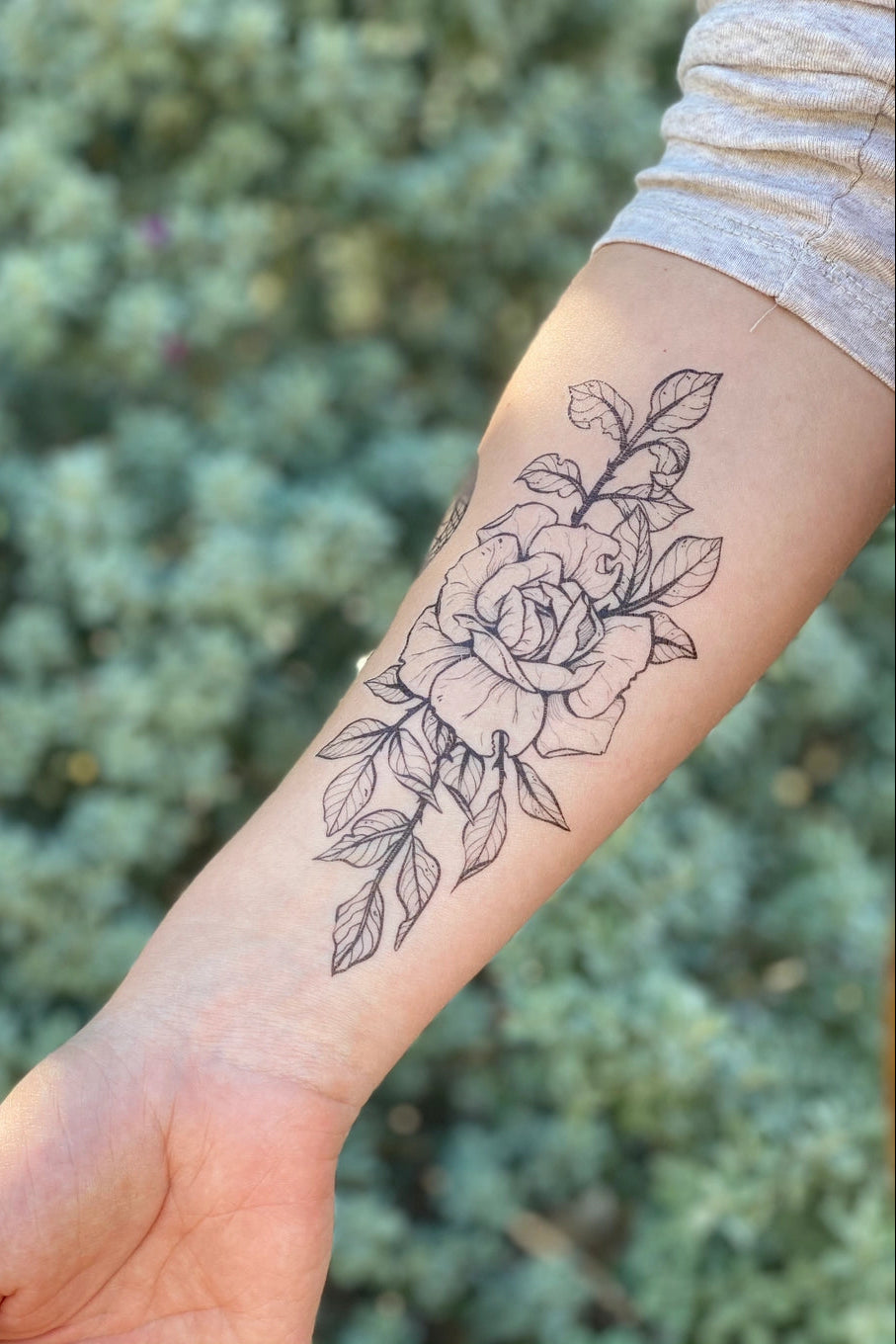 Line art rose tattoo on a person's arm with a blurred green background ©Nature Tats